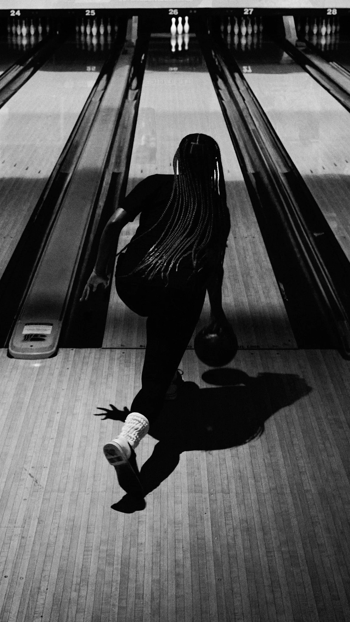 A woman with long braided hair is bowling in a bowling alley, holding a bowling ball, with her shadow cast on the wooden lane.