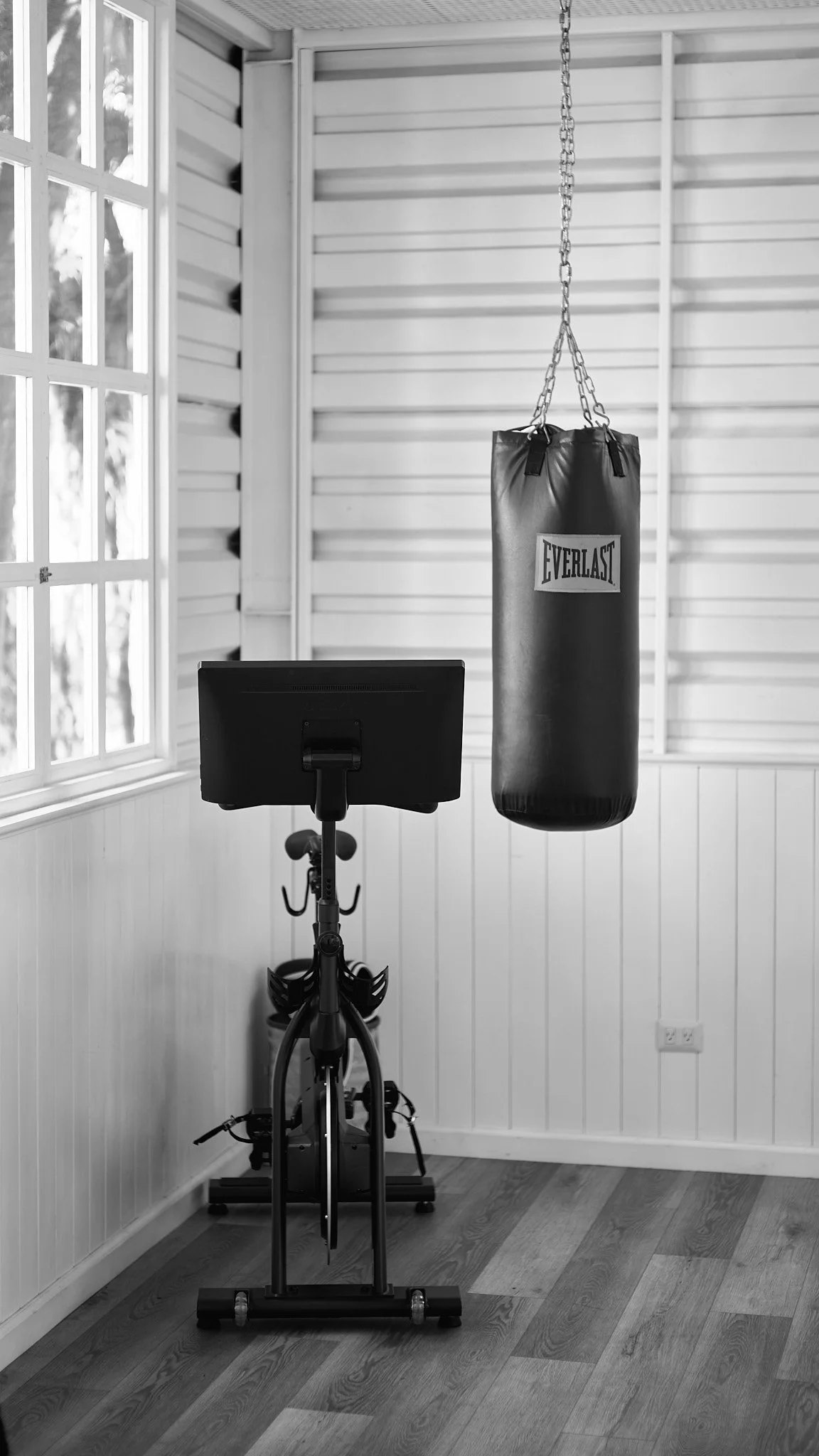 An indoor exercise corner with a bicycle, a punching bag hanging from the ceiling, and wooden flooring near a window with glass panes.