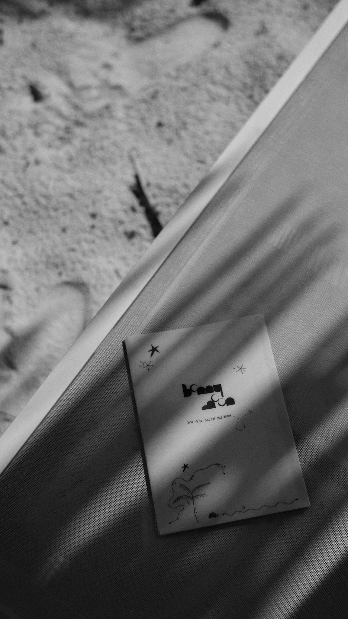 A black and white photo of a paper with illustrative designs resting on a surface, with sand in the background.