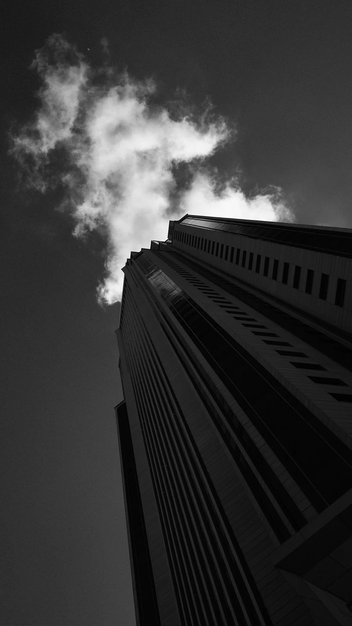 Low-angle black and white photo of a tall skyscraper with vertical lines, clouds in the sky above.