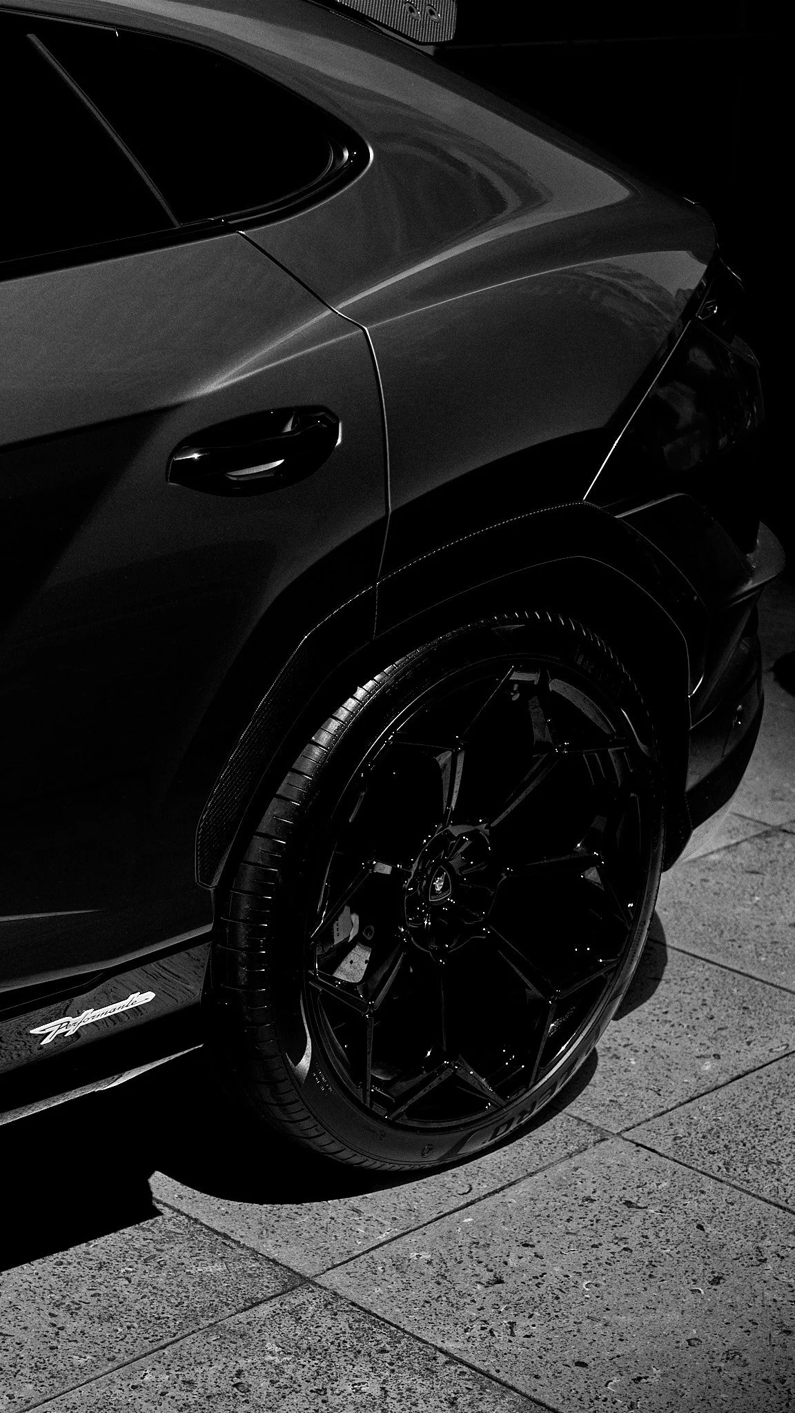 Close-up of a luxury black sports car parked on pavement, showing the rear wheel, part of the car's rear, and a sleek design.