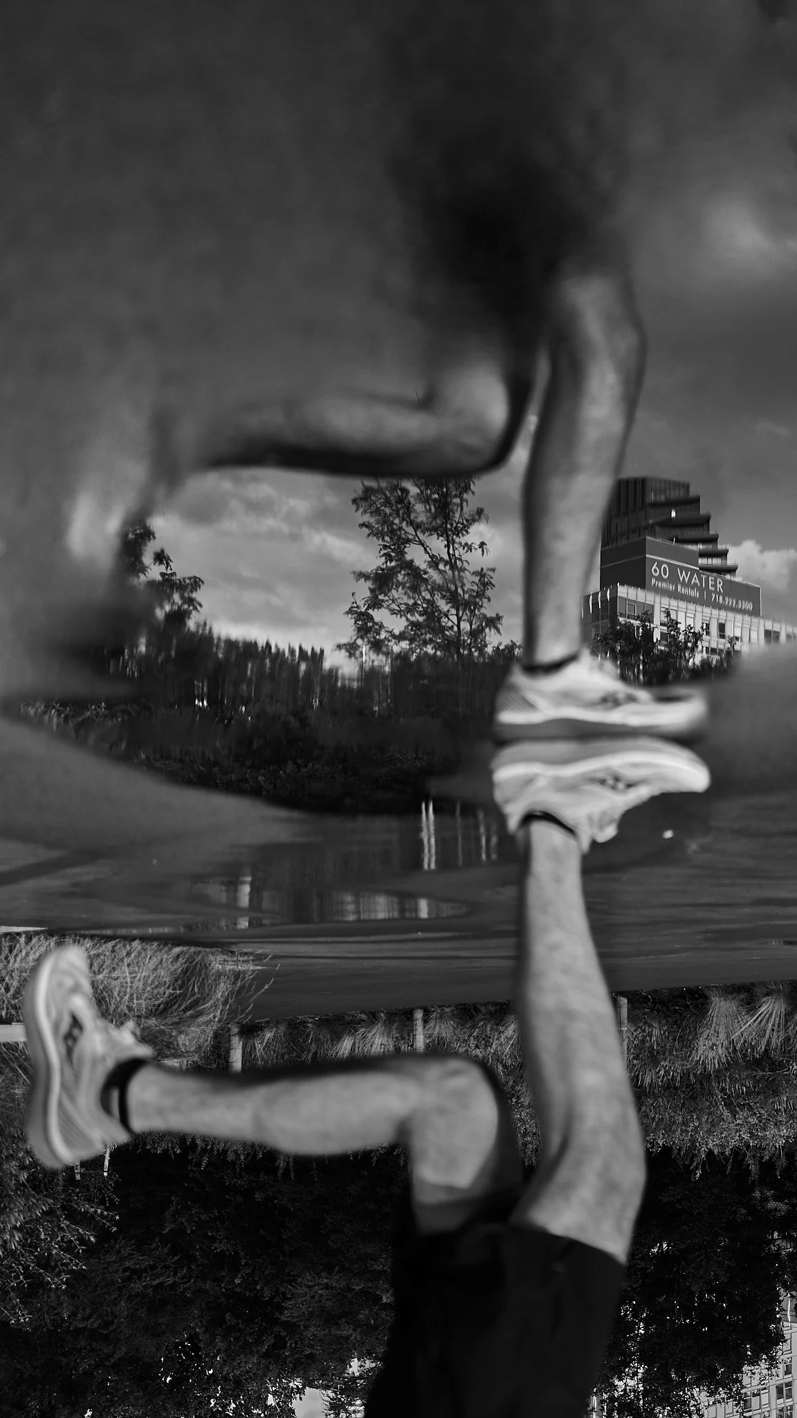 Black and white photo of a person walking through water in an urban park, with trees and a modern building in the background.