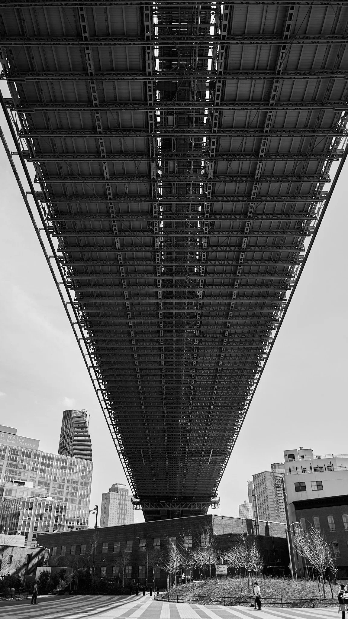A black and white photo of an underside view of a large bridge with steel girders and structure, spanning over a cityscape with modern buildings and a few trees and pedestrians below.