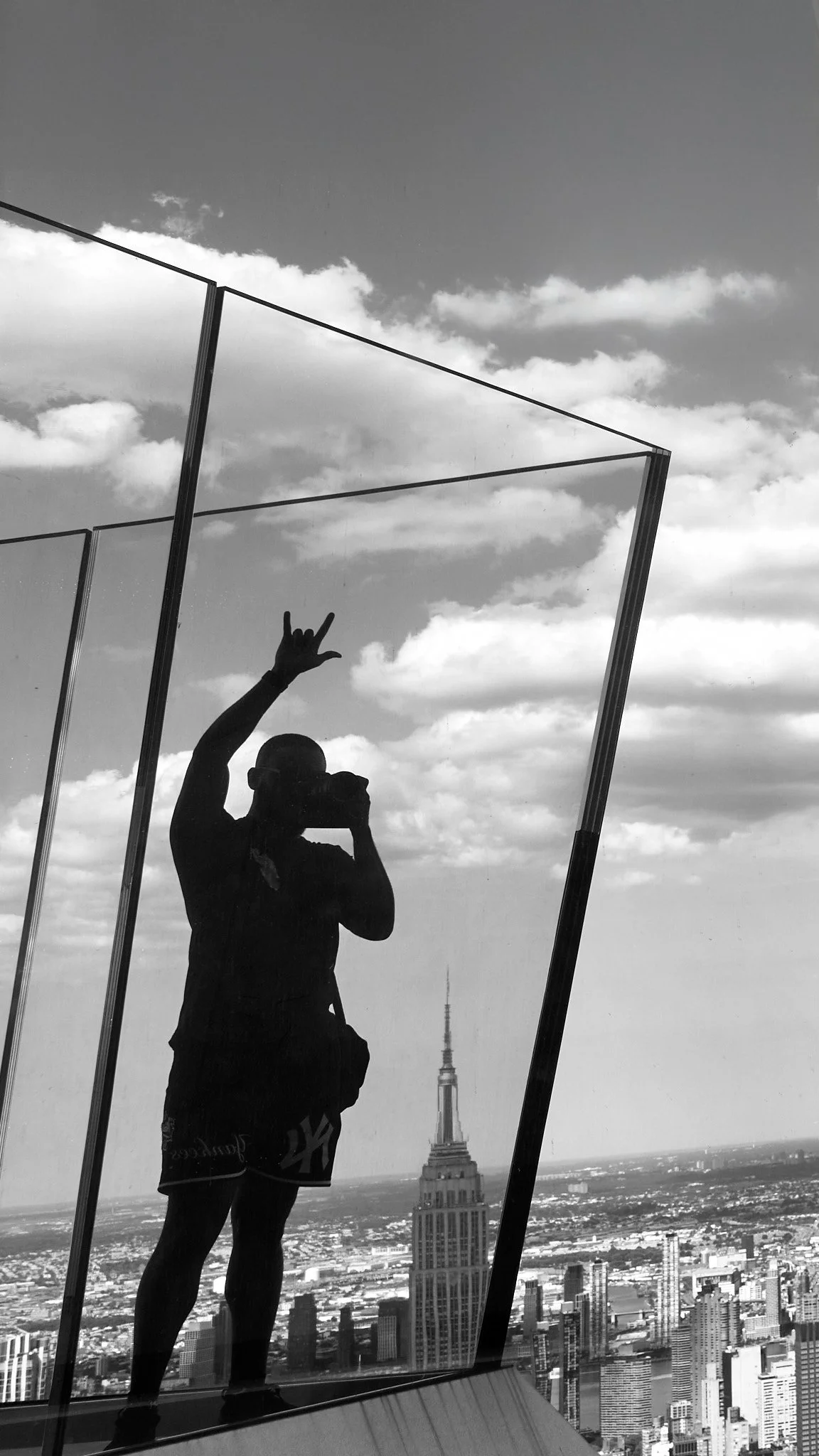 A silhouette of a person taking a selfie in front of a mirror on a rooftop with the New York City skyline, including the Empire State Building, in the background.