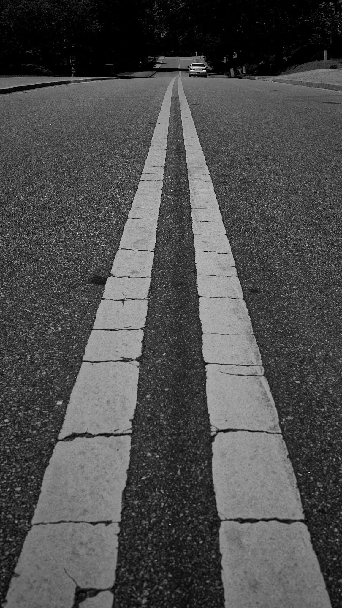 Black and white photo of a street with double yellow lines in the center, ending at a white car in the distance, with trees and houses on the sides.