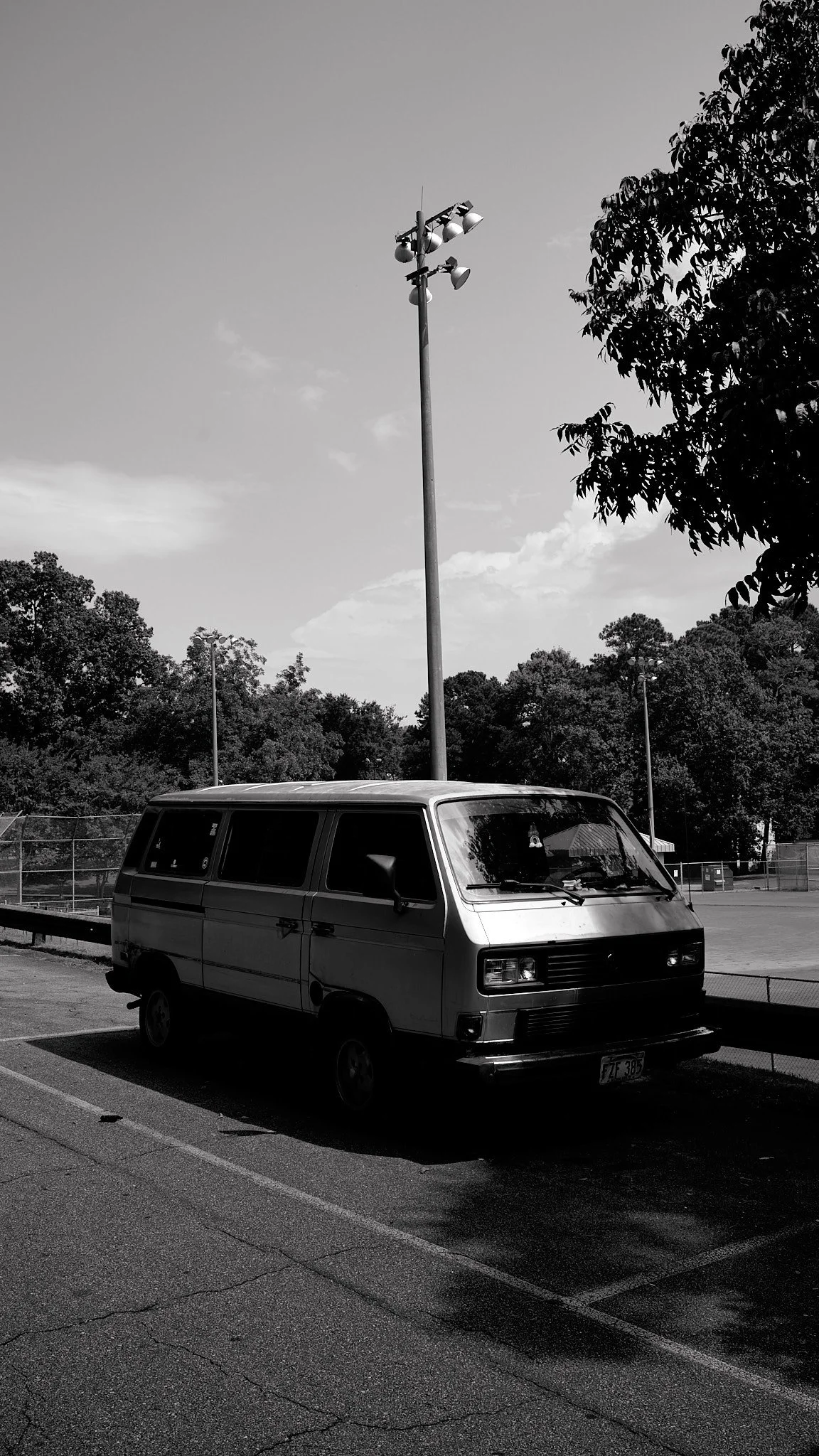 A vintage minivan parked in a parking lot with a light pole and trees in the background, captured in black and white.