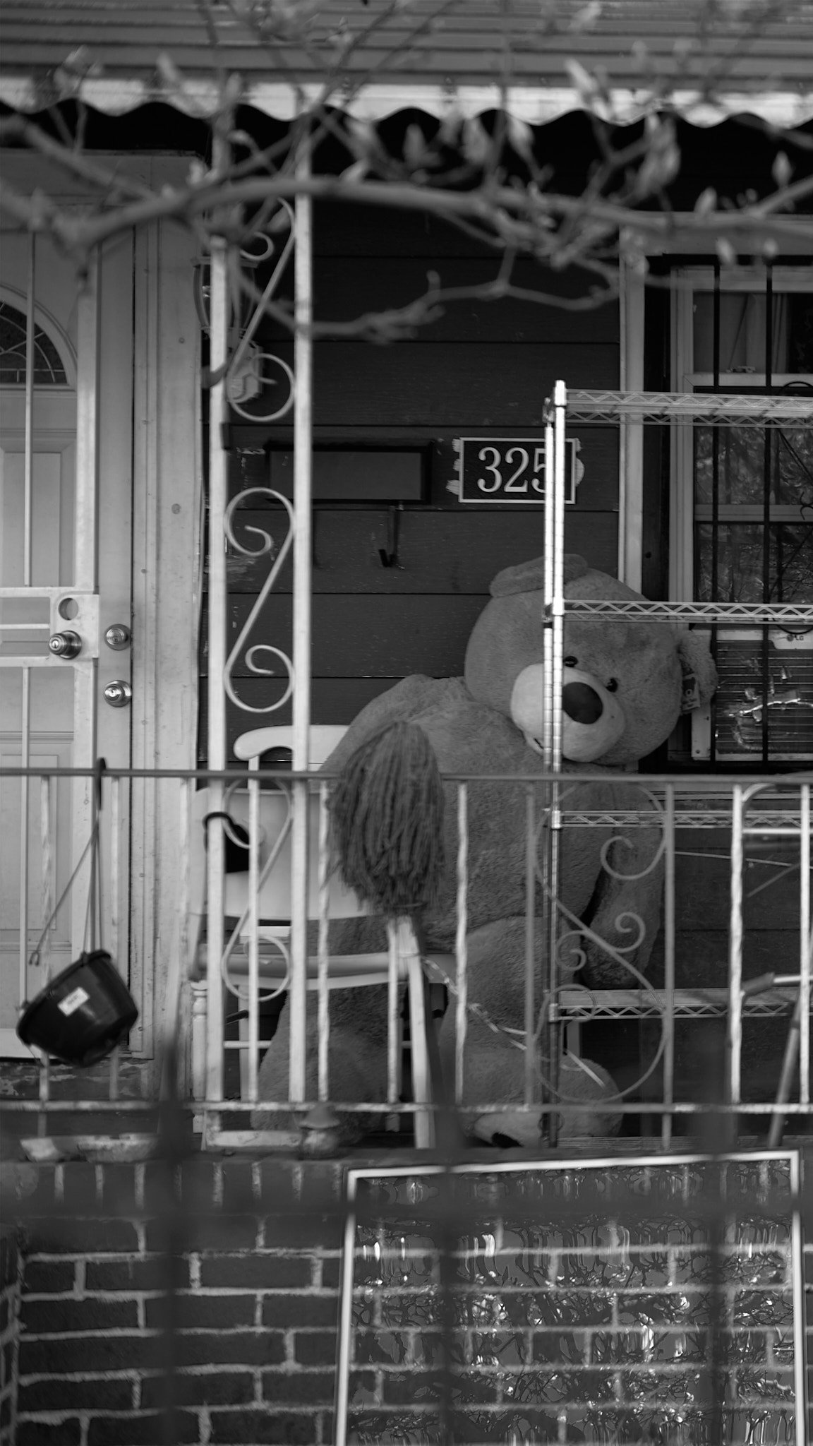 A large teddy bear sitting on the porch of a house behind a metal fence.