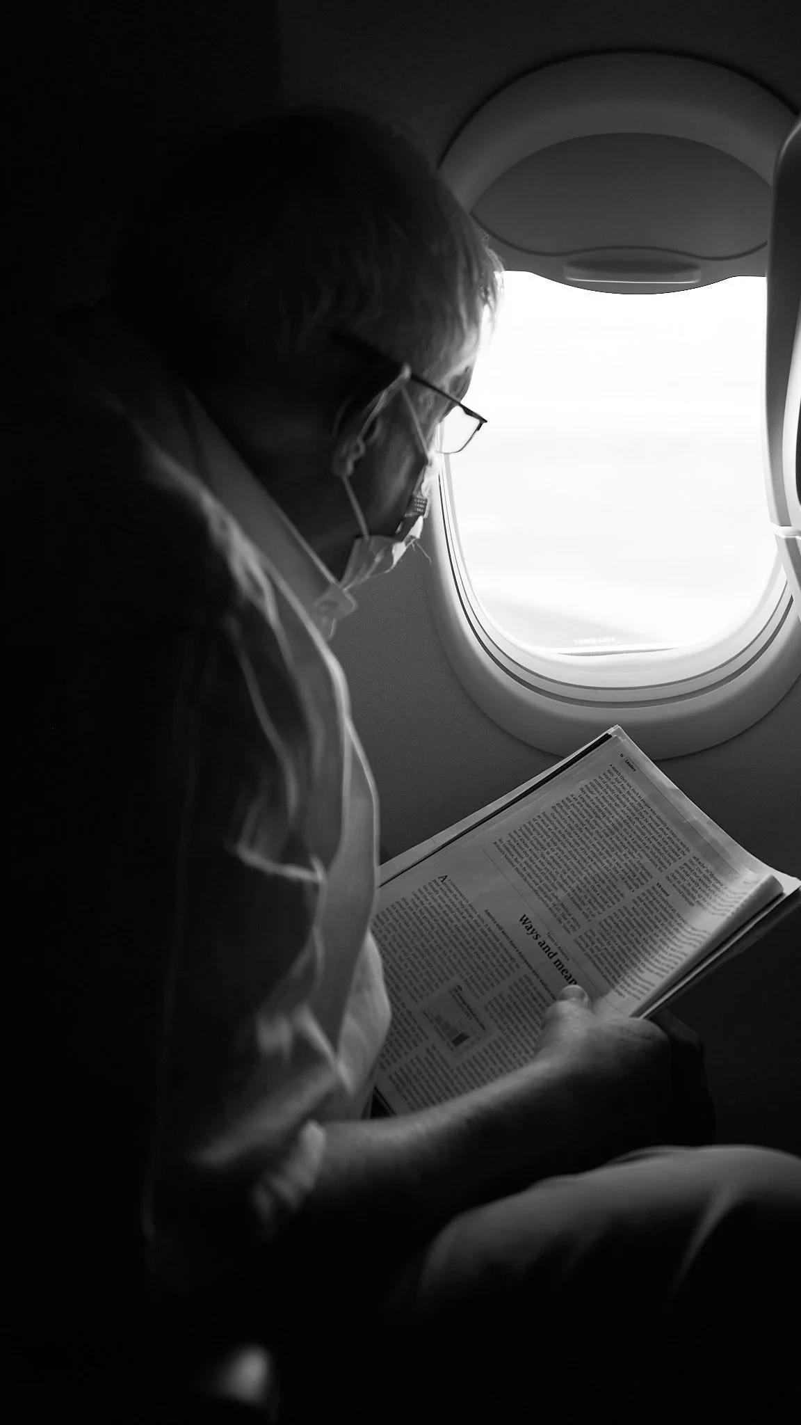 A person wearing glasses and a face mask, sitting on an airplane seat, reading a newspaper, with airplane window in the background.