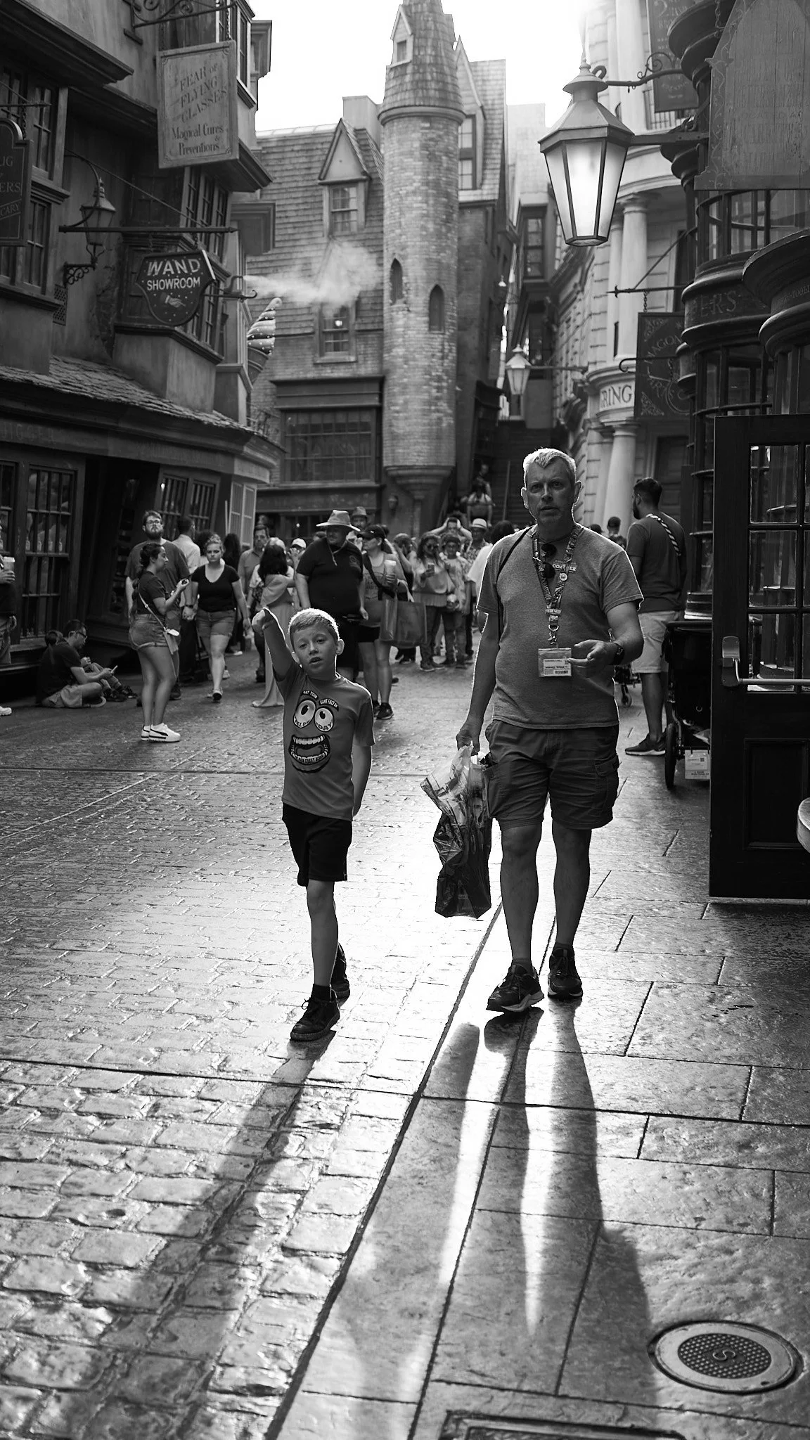 A black and white photo showing a busy street scene with many people walking, some sitting, in front of buildings that resemble a fantasy or storybook setting, with towers and whimsical architecture. A young boy walks beside an older man carrying a s