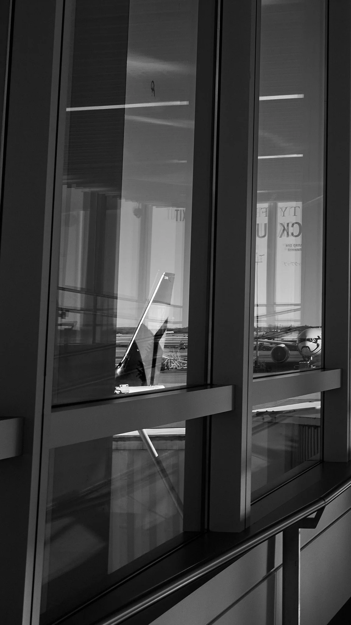 View of an airport terminal interior with glass walls, showing reflections and an airplane parked outside, captured in black and white.