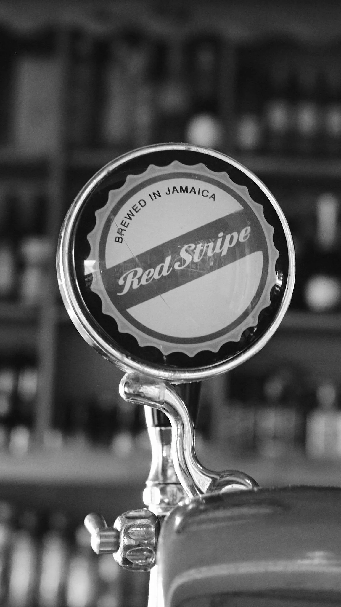 Close-up of a keg tap handle labeled "Red Stripe," indicating it is brewed in Jamaica, with a blurred background of a bar or brewery.