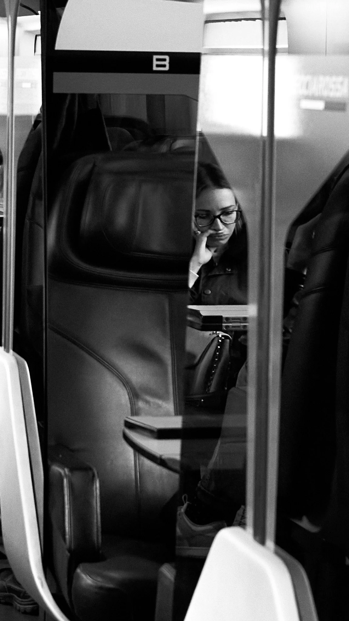 A woman with glasses is sitting at a desk inside a bus, resting her chin on her hand and looking down at some papers or a book.