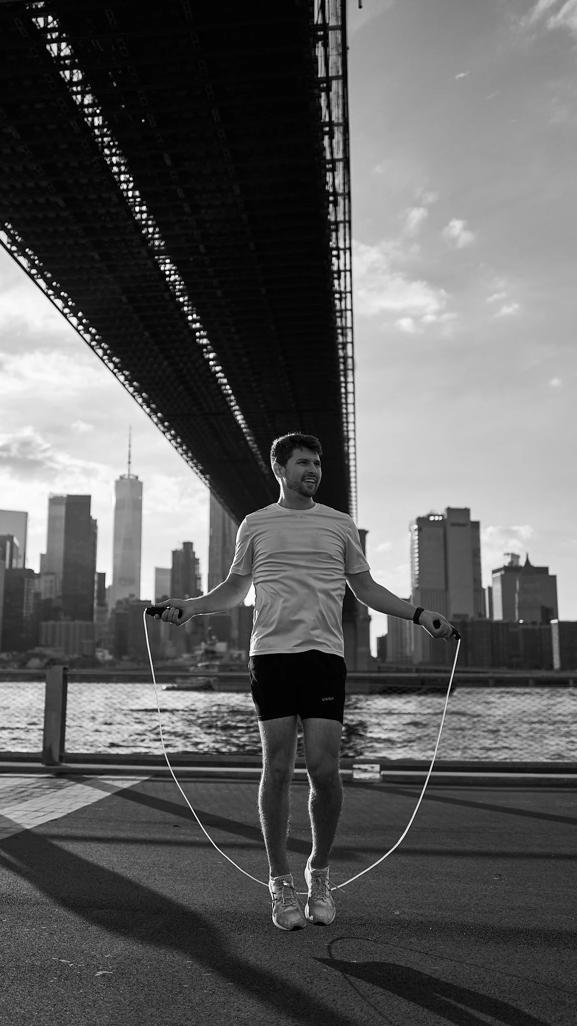 A man exercising with a jump rope under a bridge in an urban city, with tall buildings and a river in the background.