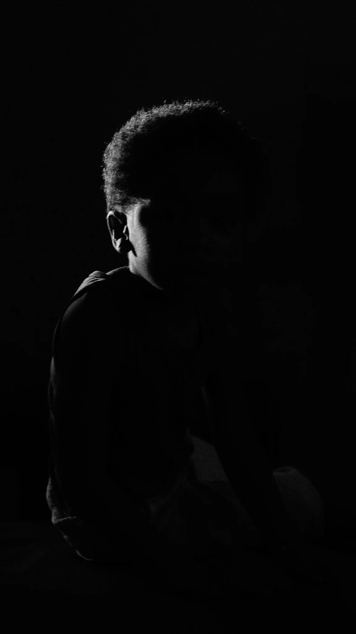 Silhouette of a boy sitting in low light, showing only the side of his head and shoulder, with a dark background.