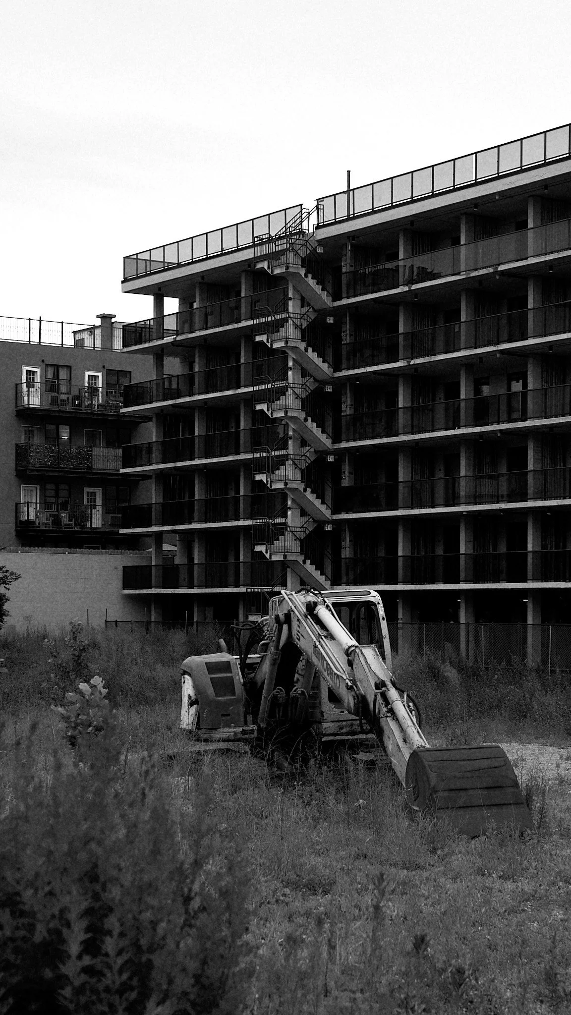 A black and white photo of a construction excavator in front of a modern apartment building with multiple levels and balconies, and external staircases.