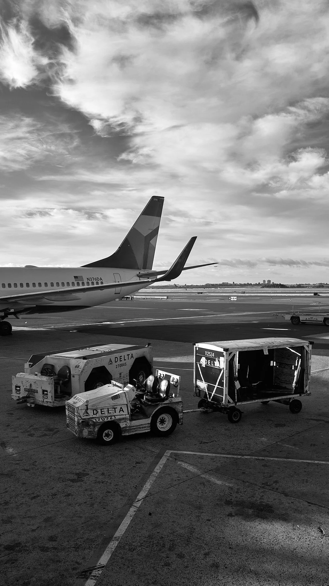 Black and white photo of a Delta airplane at an airport, with luggage carts, baggage tow tractor, and a cloudy sky in the background.