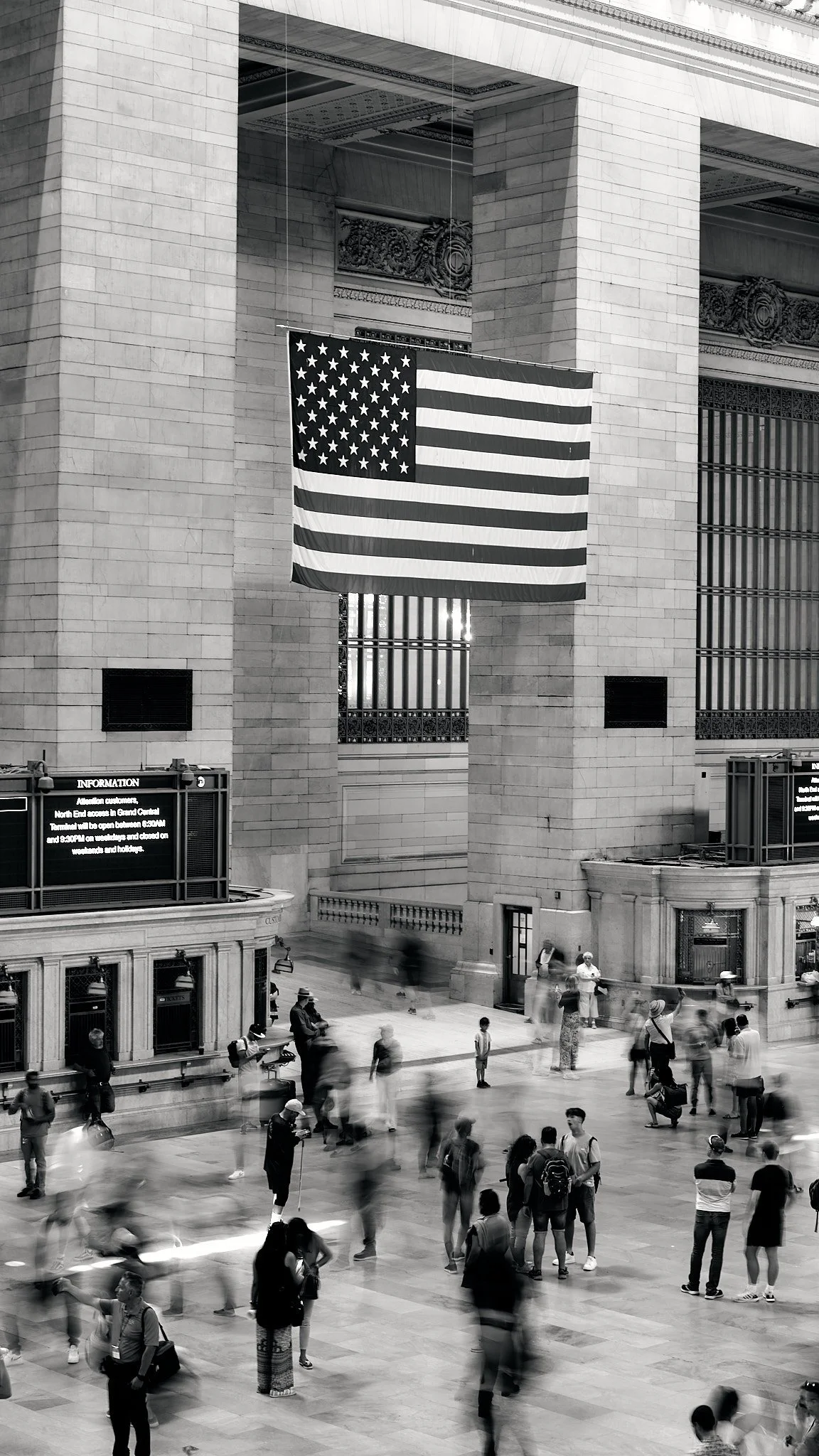A large American flag hangs inside a grand, historic train station with stone columns and high ceilings. People are walking and waiting in the busy station, some blurred due to motion.