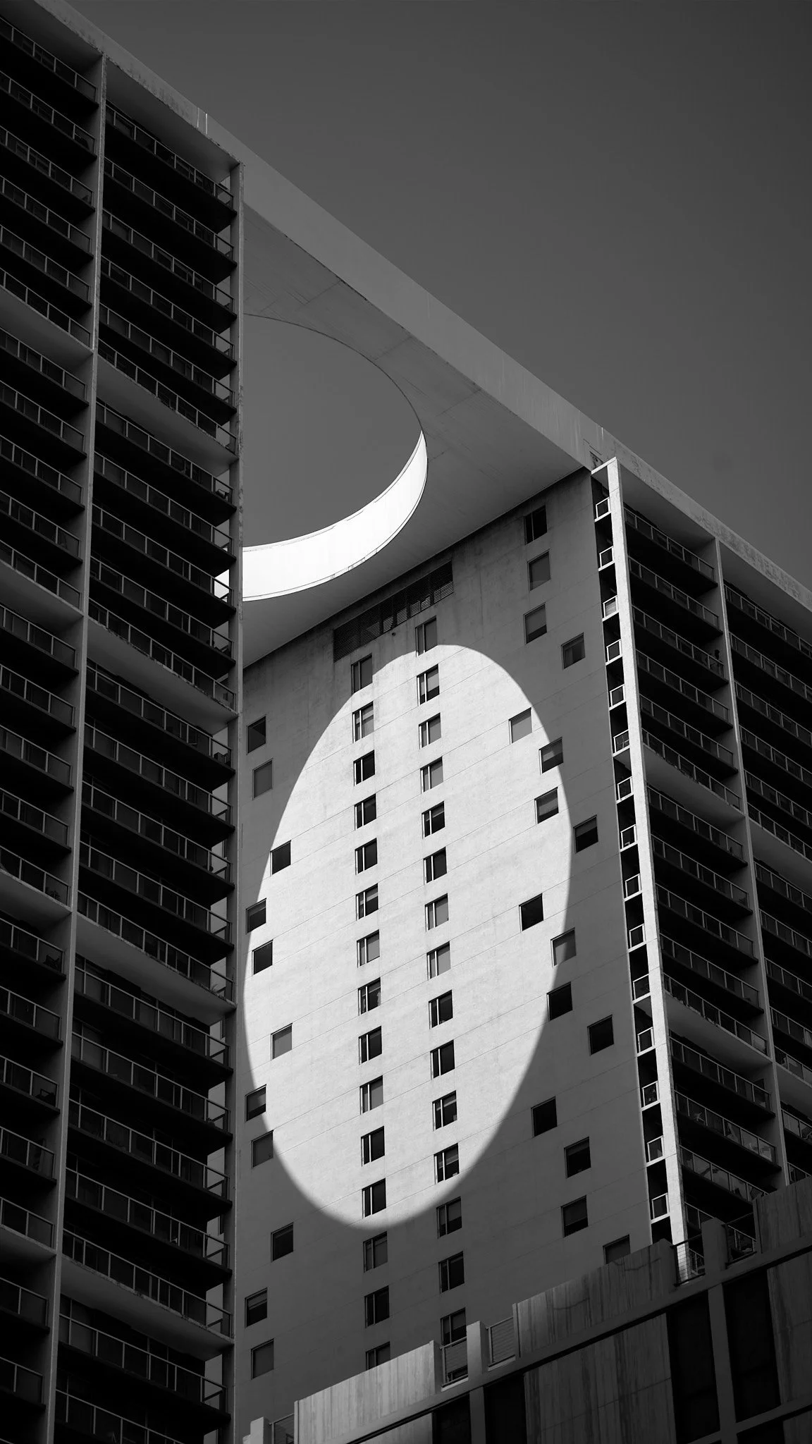 Black and white photo of modern high-rise building with circular opening and oval-shaped shadow on the building's facade.