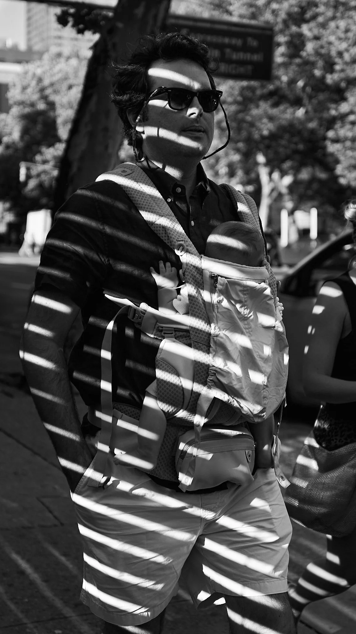 A person wearing sunglasses and a backpack carrying a baby in a front-facing carrier, standing outdoors on a sunny day with tree shadows.