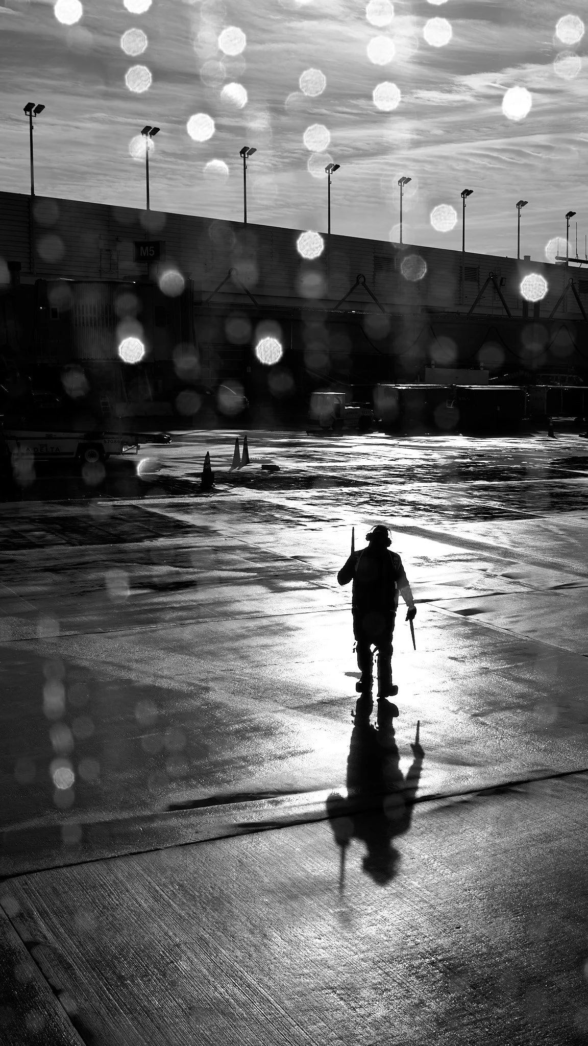 A person walking on a wet airport tarmac with rain droplets on the camera lens, silhouetted against a bright sky with clouds, airport buildings, and parked vehicles in the background.