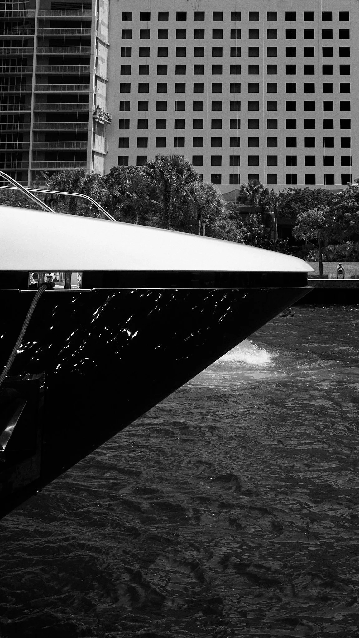 Close-up of a yacht docked in a waterway with modern buildings and trees in the background, all in black and white.