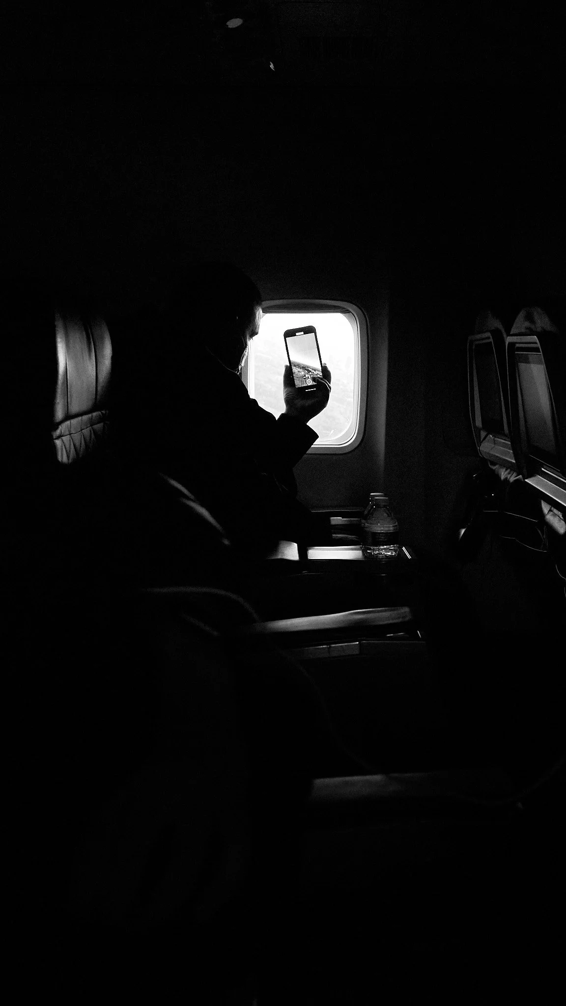 Passenger sitting on airplane window seat, taking a photo outside with their phone, water bottle on tray table, dark interior with overhead lights off.