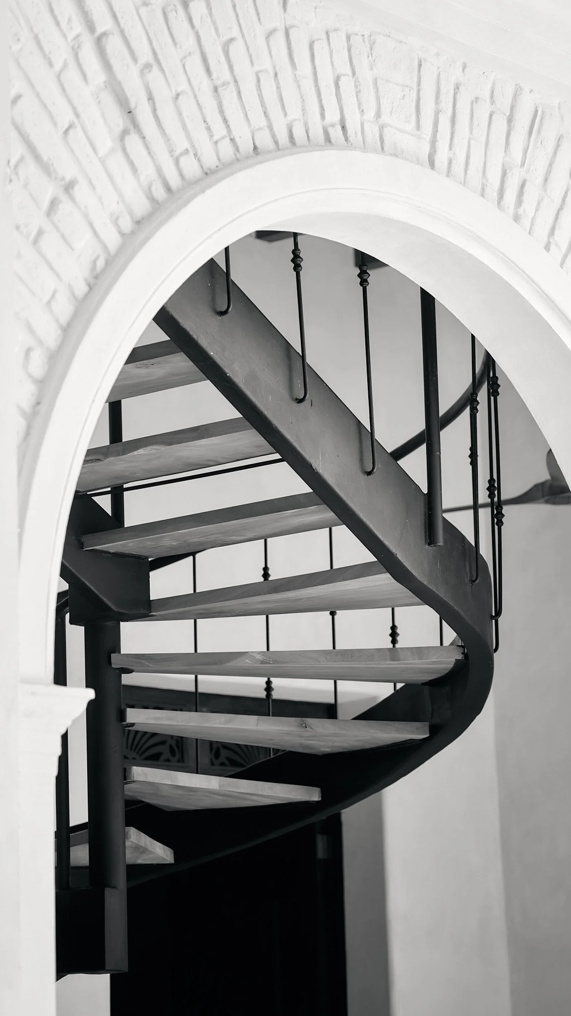 Black and white photo of a spiral staircase seen through an arched opening, with wooden steps and metal railing with vertical balusters, against a light-colored wall.