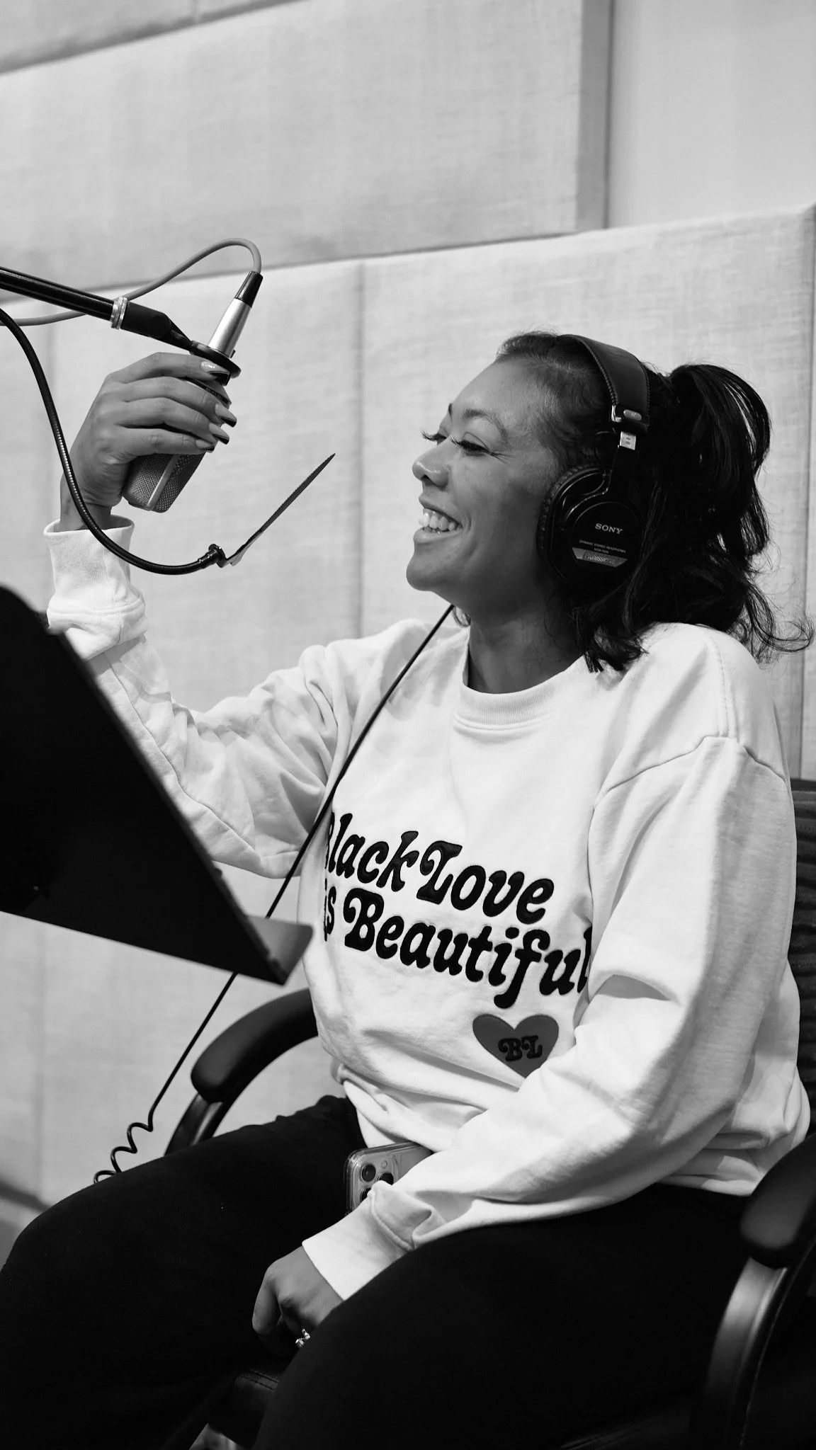 A woman in a recording studio wearing headphones, singing into a microphone, and smiling. She is wearing a sweatshirt that says 'Black Love is Beautiful'.