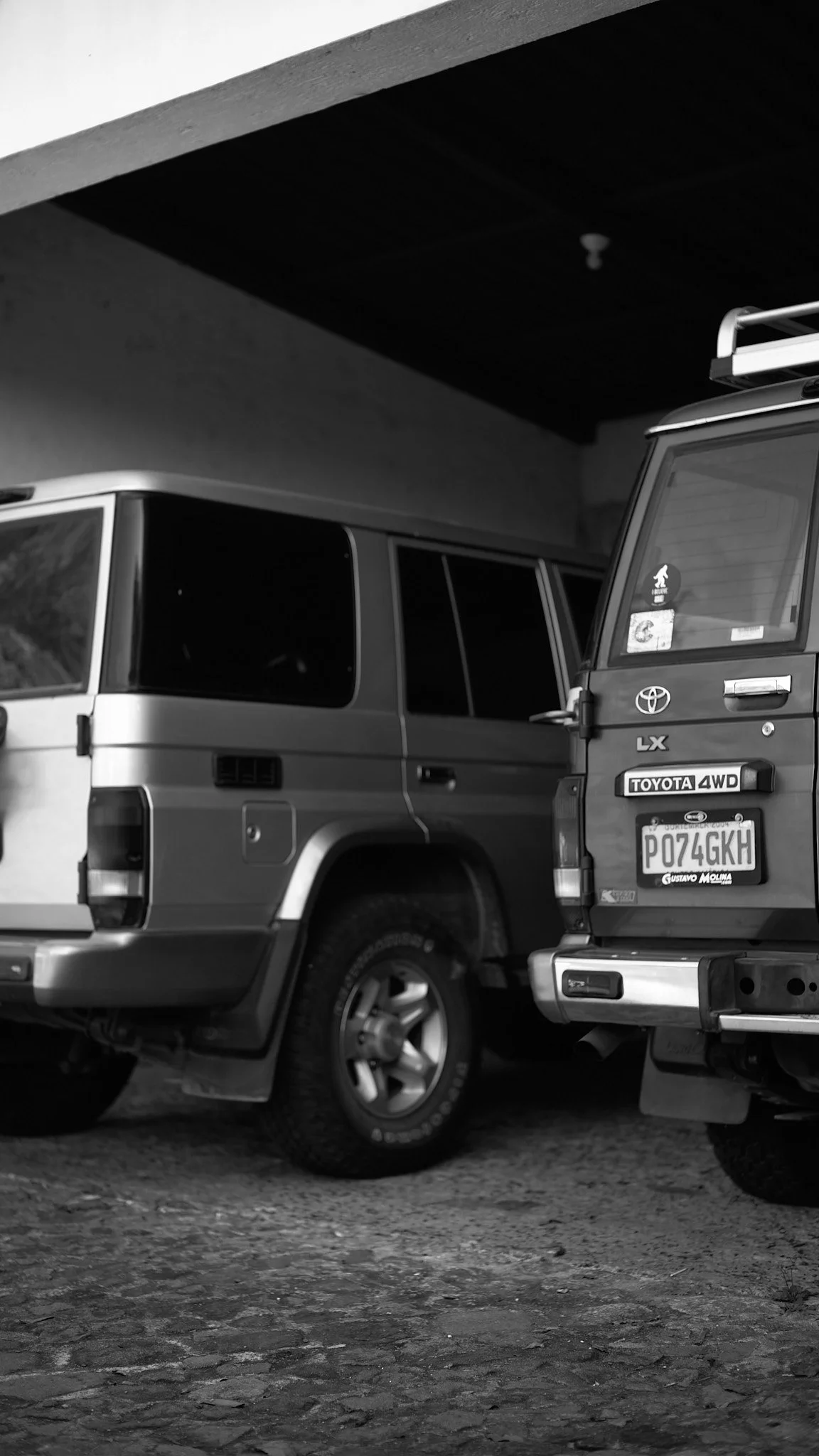 Two parked Toyota SUVs, one behind the other, in a garage with a rough stone floor and a ceiling with a visible light fixture, in black and white.