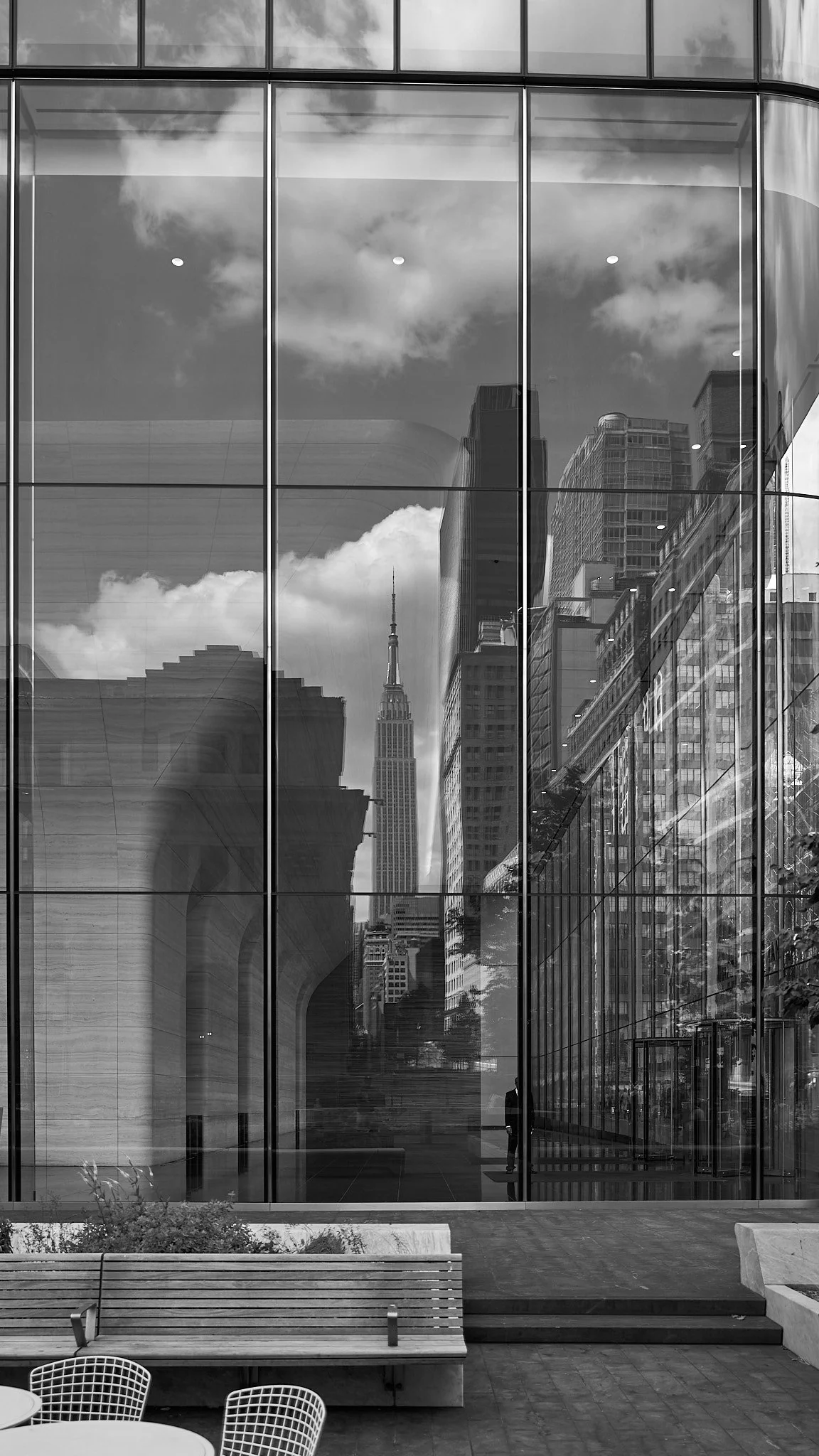 Reflective glass building exterior with the Empire State Building visible in the reflection, outdoor seating area with benches and tables in front.