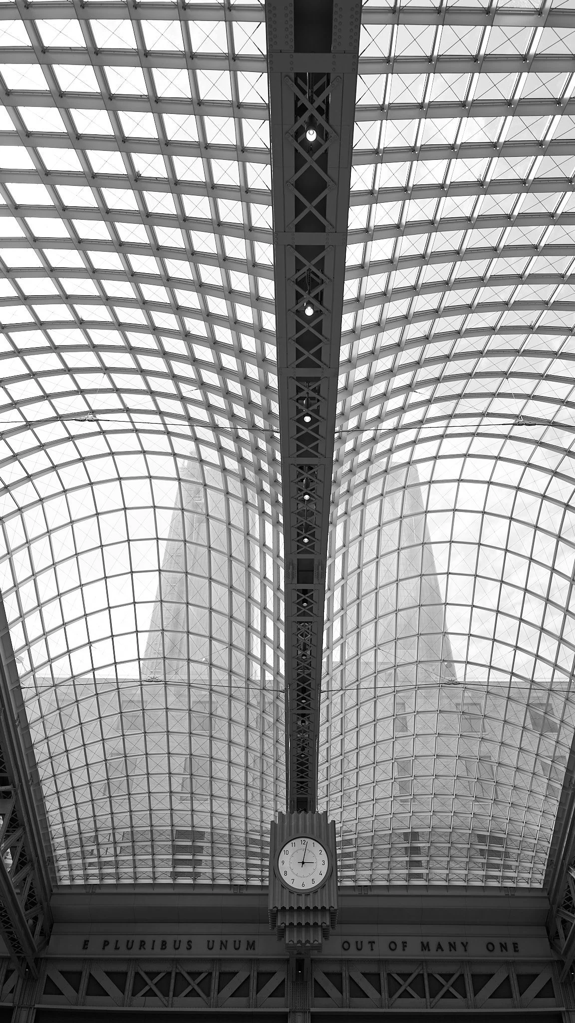 Interior view of a large glass and metal-domed ceiling with a central clock showing 12:01, and an inscribed phrase below reading 'E PLURIBUS UNUM OUT OF MANY ONE'.
