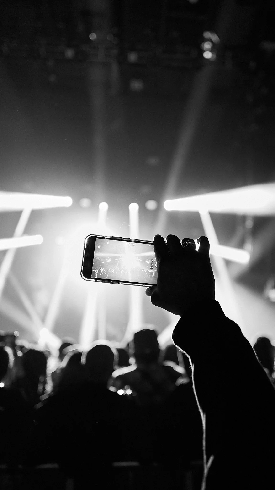 A person is holding a smartphone and taking a photo or video at a concert with bright stage lights and a crowd of people in the background, in black and white.