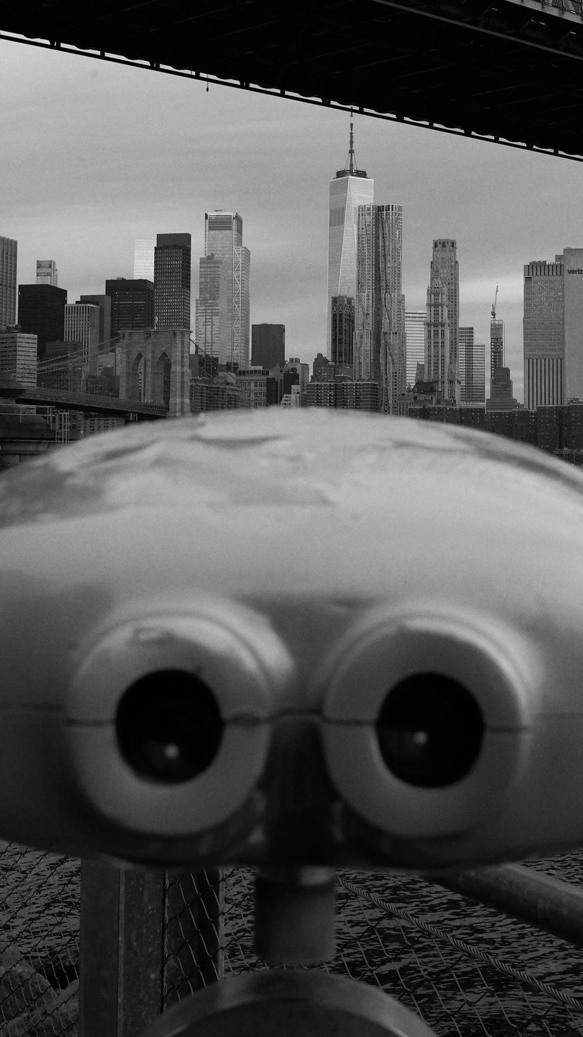 A black and white photo taken from a high vantage point, showing a city skyline with tall skyscrapers, including One World Trade Center, in the background. In the foreground, a coin-operated binocular viewer is visible, slightly out of focus, along w