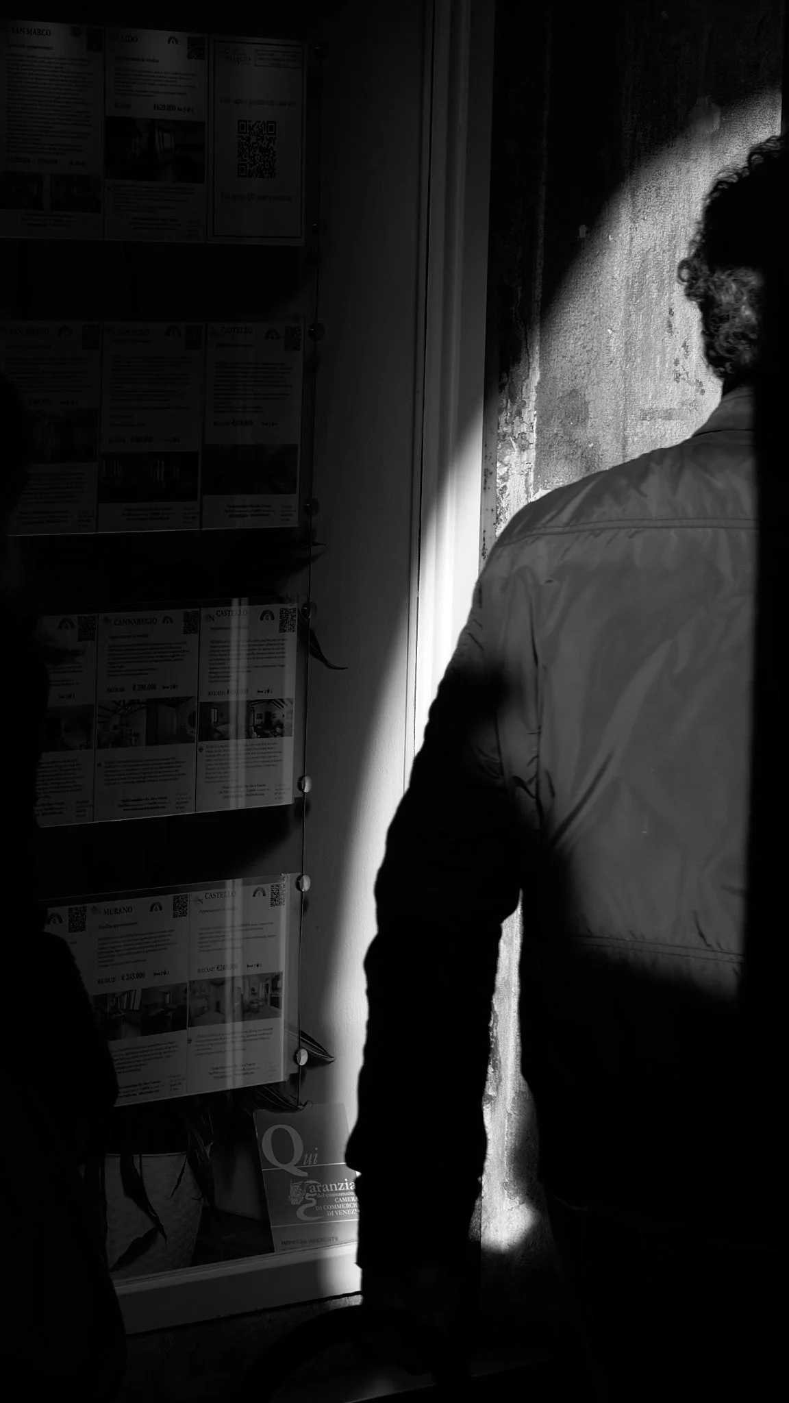 Silhouettes of two people looking at real estate listings or advertisements displayed on a wall, with a bright light illuminating the edge of a wall and casting shadows.
