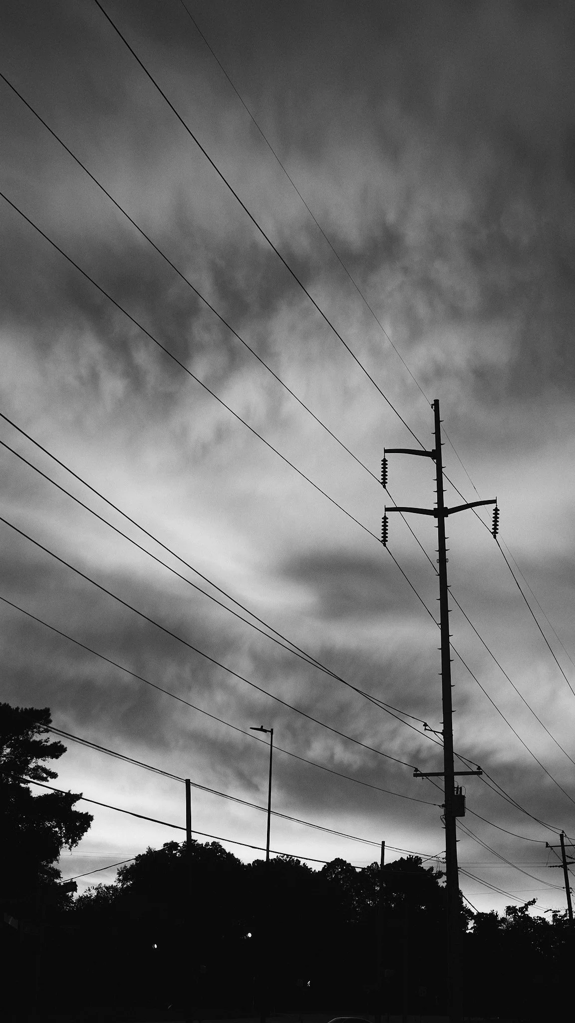Black and white photo of power lines and utility poles against a cloudy sky, with trees and streetlights in the foreground.