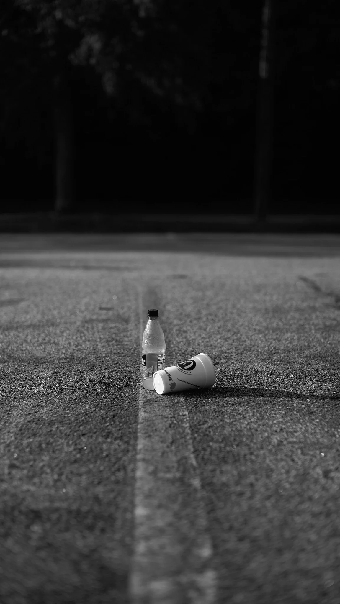 A dark street scene with two abandoned beverage containers, a plastic bottle and a paper cup, both on the ground in the foreground.