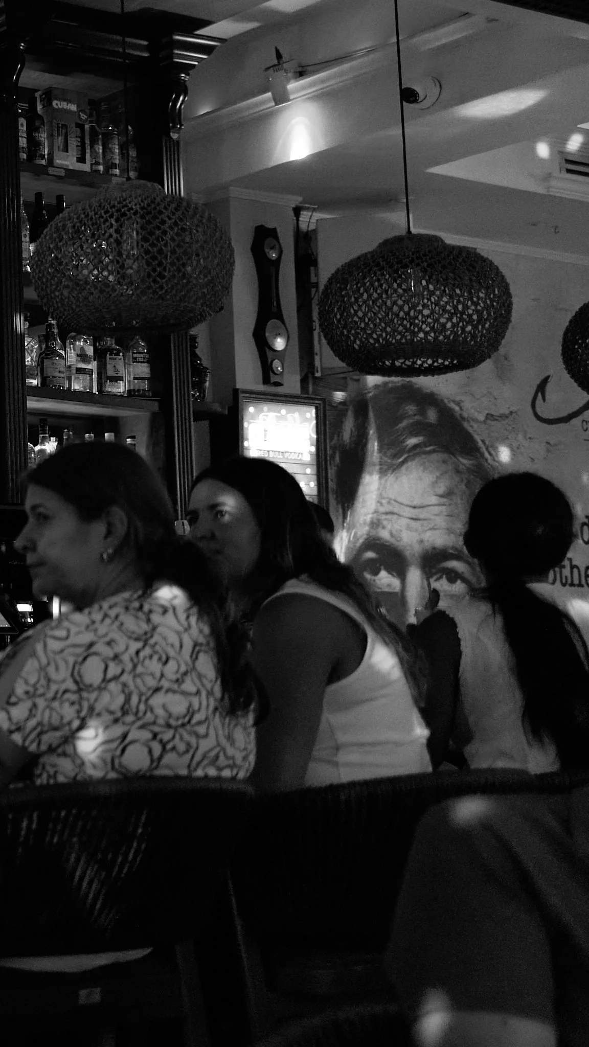 People sitting at a bar with bottles on shelves, large pendant lights, a mural of a person's face, and a wall clock in the background.