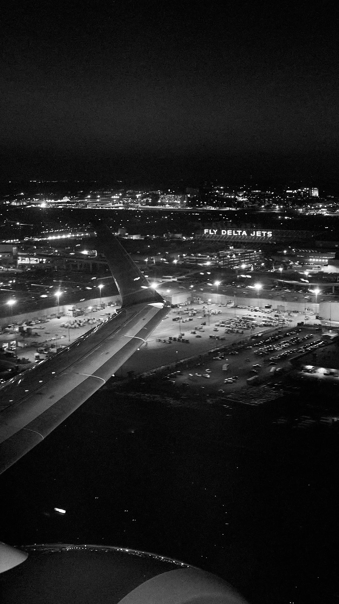Nighttime aerial view from an airplane window showing the airplane wing, airport grounds with parked planes and vehicles, and a sign reading 'Fly Delta Jets' in the distance.