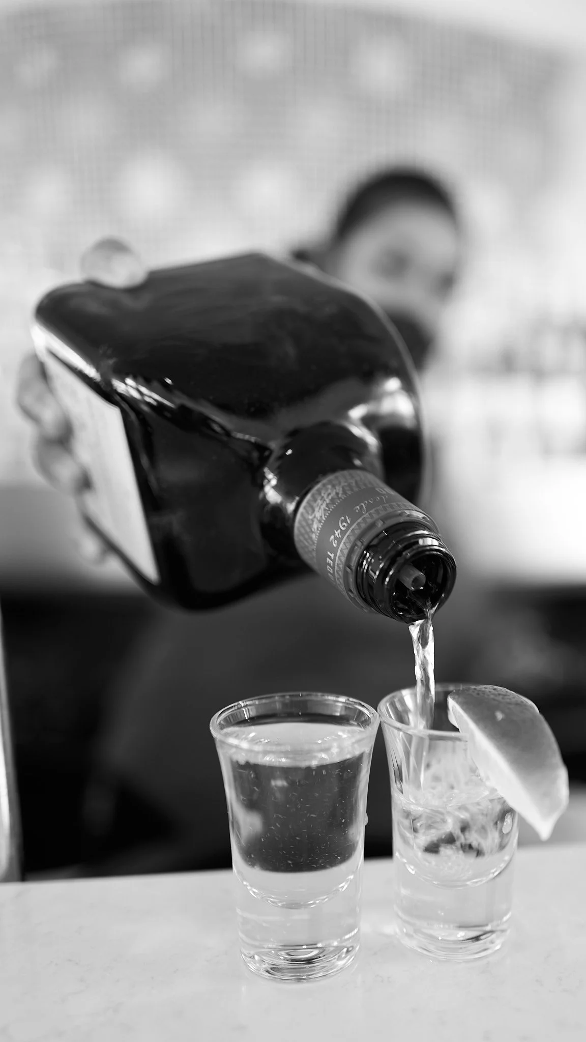 A person pouring a bottle of tequila into a shot glass with a lemon wedge on the glass, black and white photo.