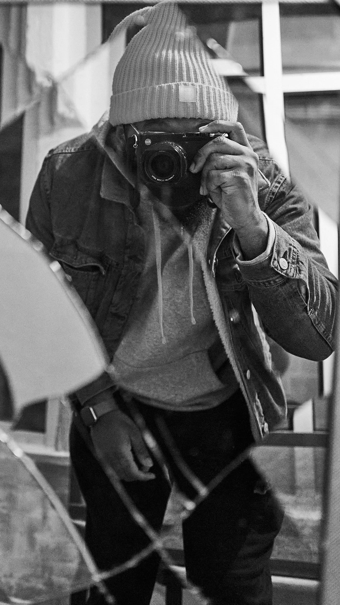 Person taking a mirror selfie with a camera, wearing a beanie, hoodie, and denim jacket in a room.