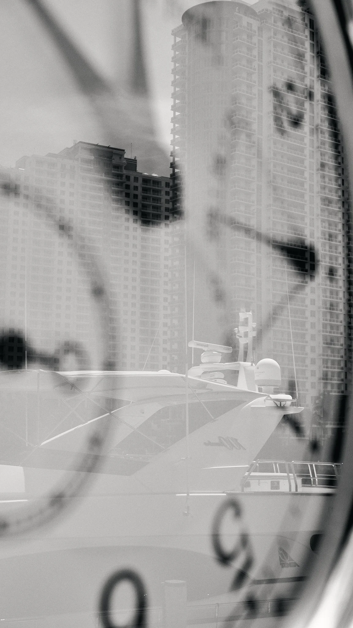 A black and white photo of a yacht docked near a city, with tall buildings in the background, seen through a window or glass surface.