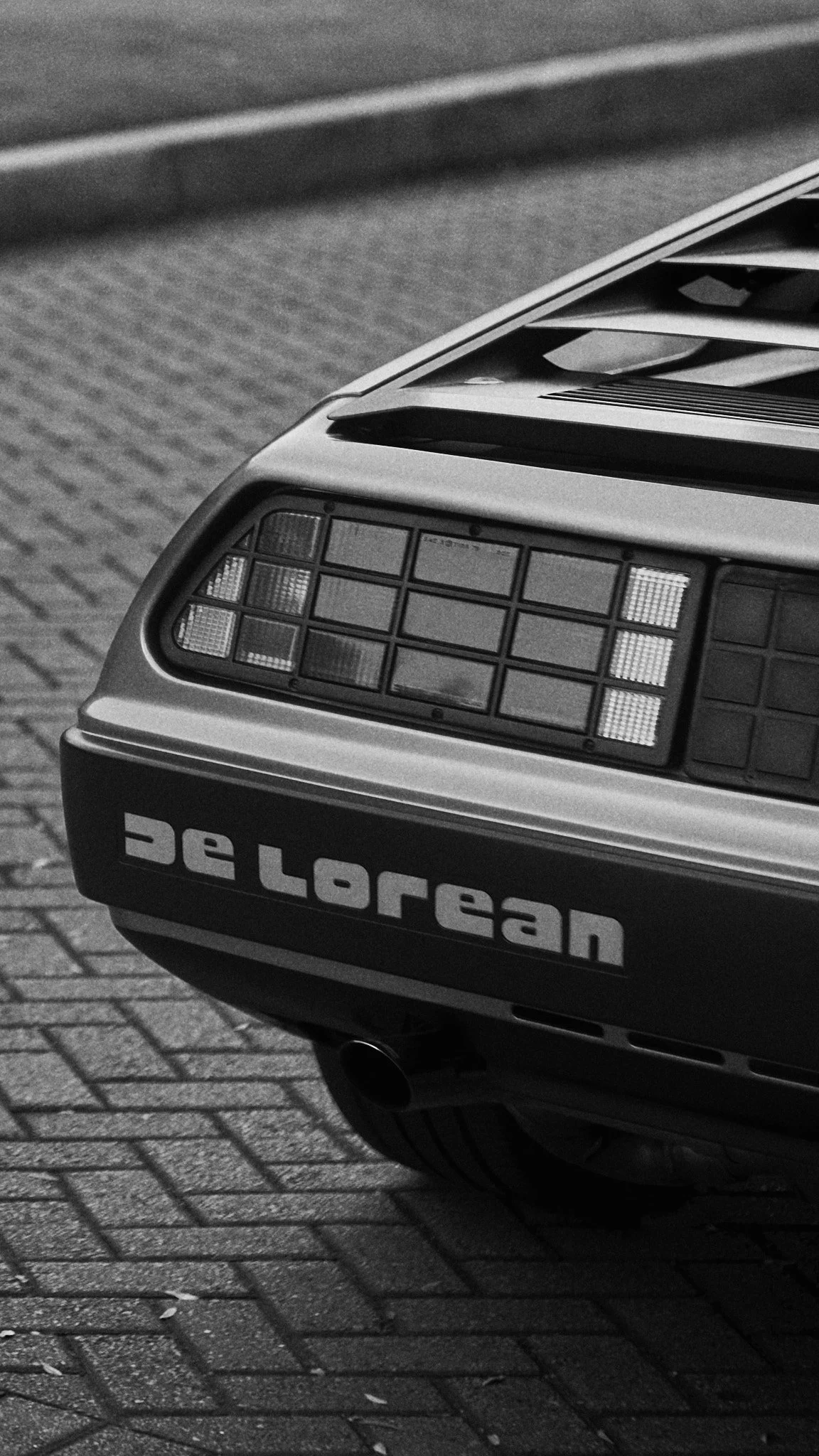 Close-up of the rear end of a DeLorean car showing the taillights, exhaust pipe, and 'deLorean' logo on the bumper, parked on a brick-paved street.