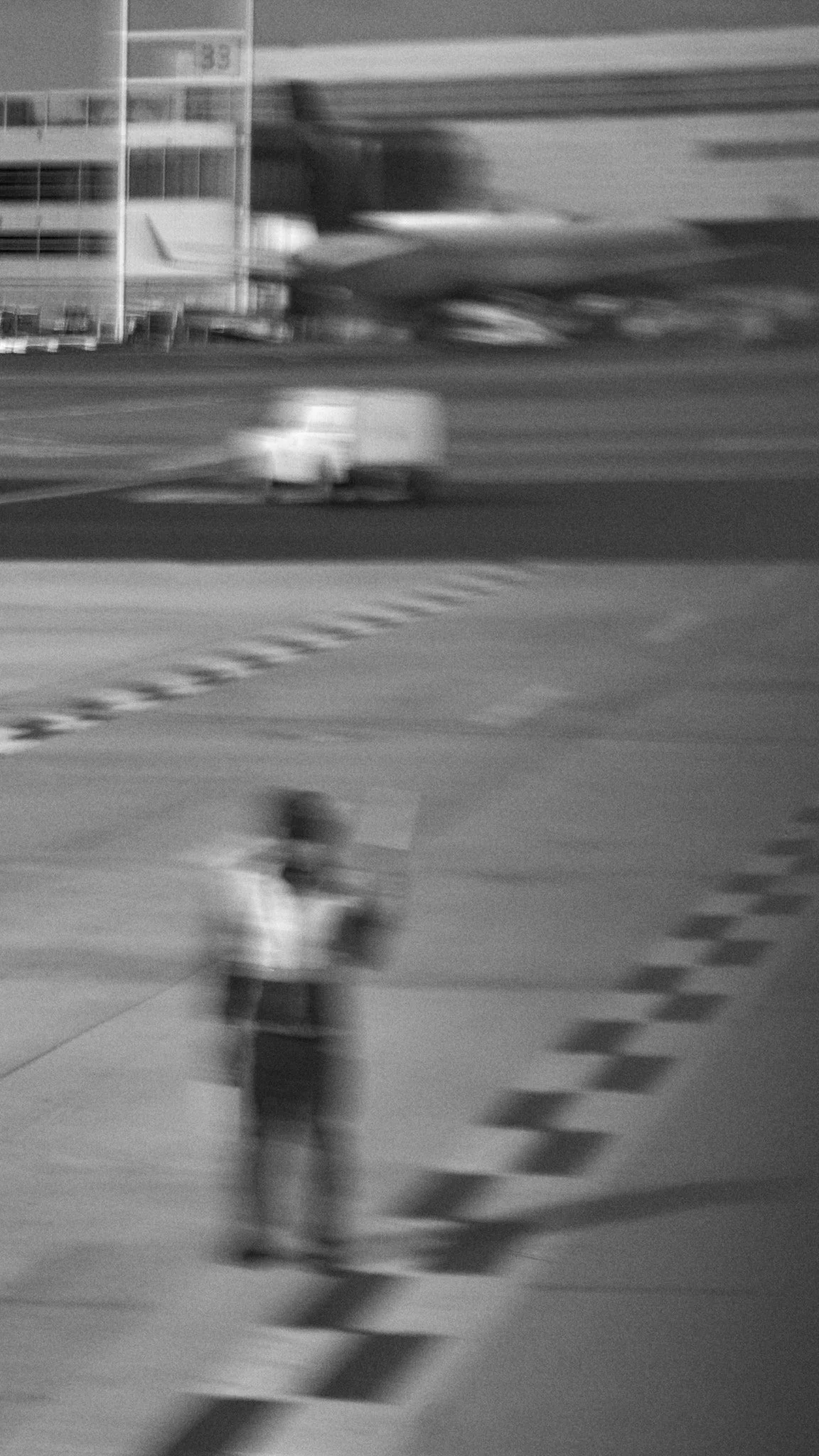A blurry black and white photo of a person walking in an airport or train station, with a large building in the background and a small vehicle in motion on the road.