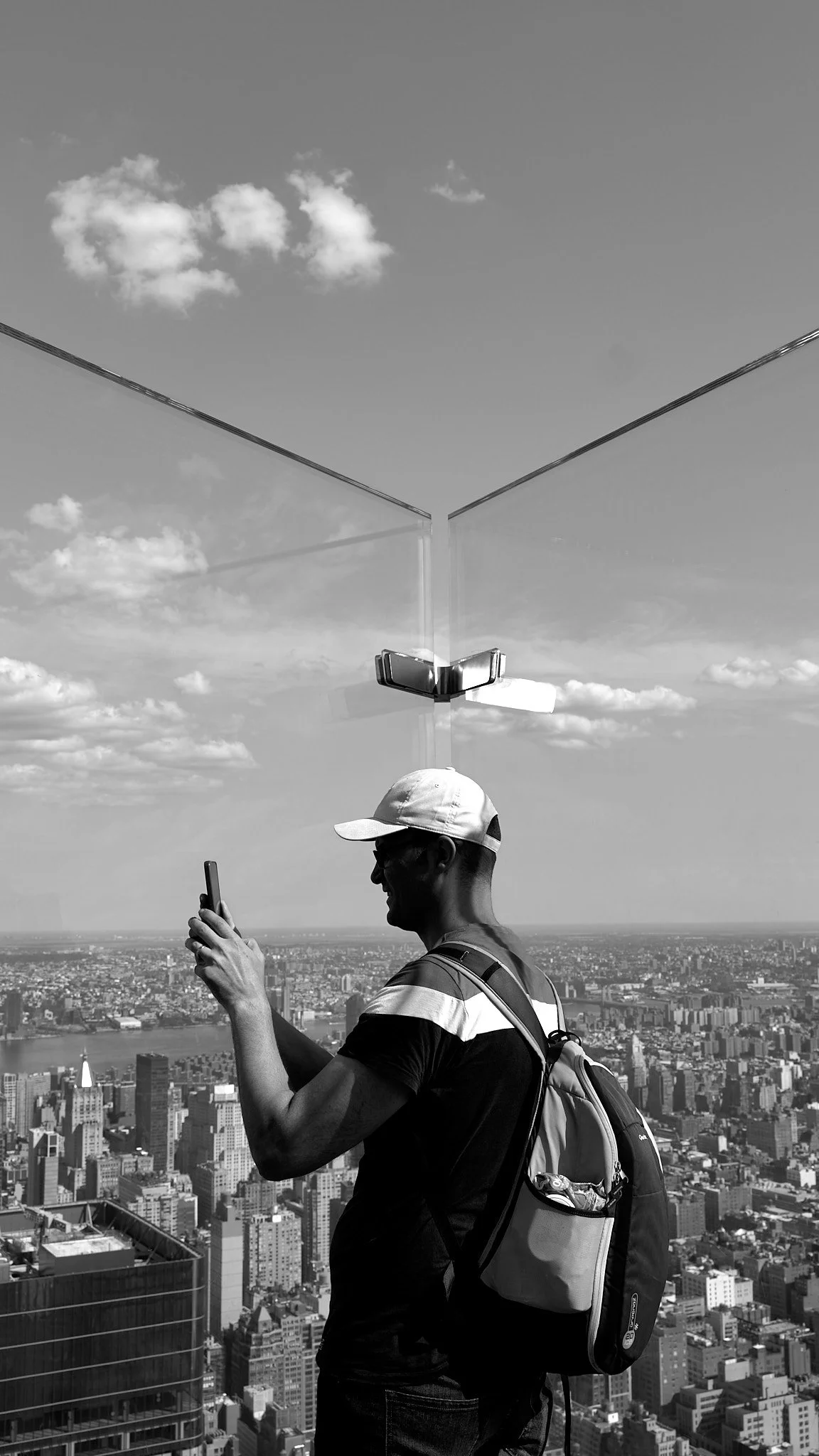 Man taking a selfie on a rooftop overlooking New York City skyline with tall buildings, river, and partly cloudy sky.