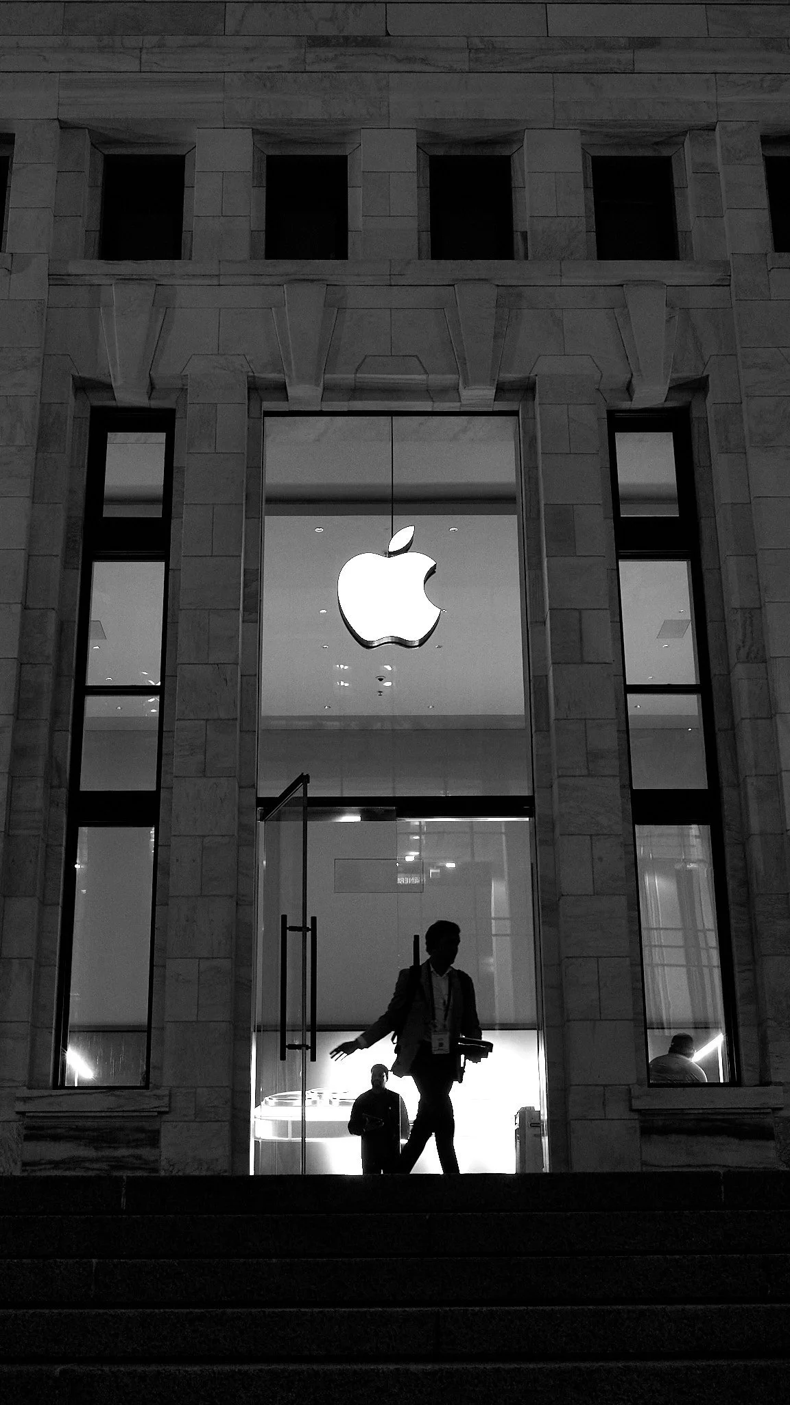 Front view of an Apple Store with a large illuminated Apple logo on the glass entrance, people walking inside, stone architecture, and a staircase leading to the store.