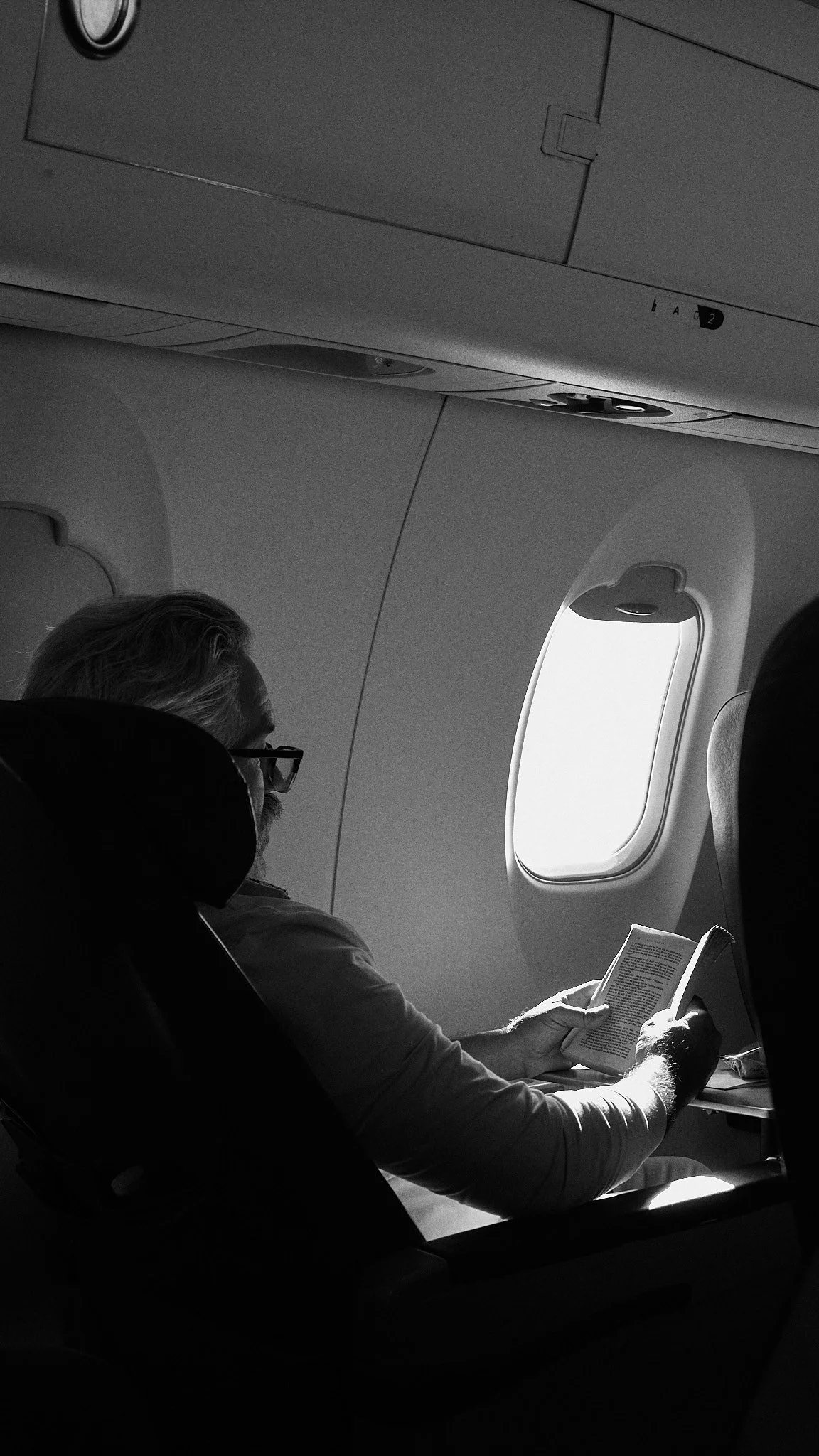 A man with glasses and gray hair reading a book while seated on an airplane near the window.