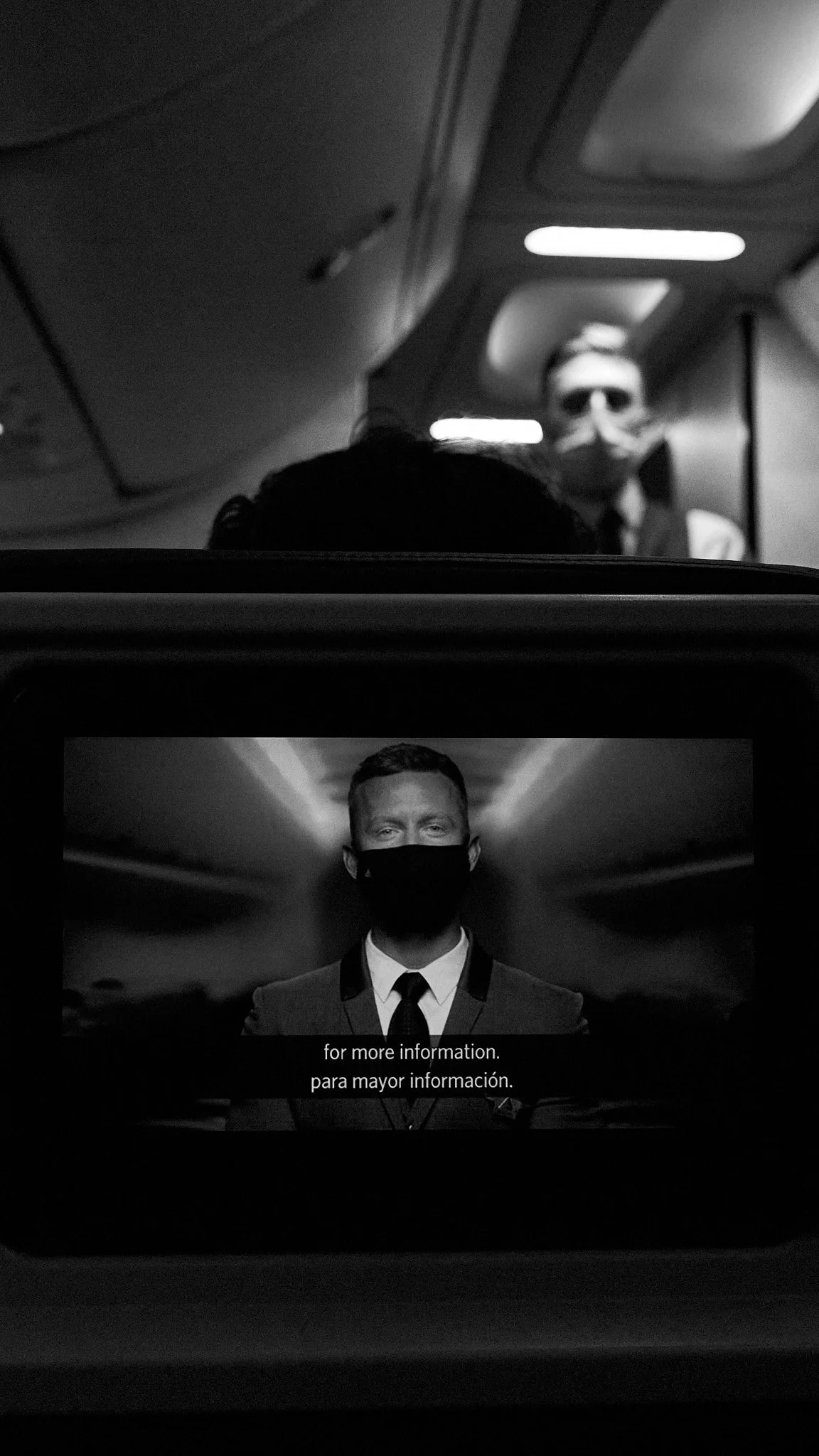 In-flight entertainment screen showing a man in a suit wearing a face mask with subtitles that read 'for more information, para mayor información.' The background appears to be a flight cabin with overhead compartments and a flight attendant wearing 