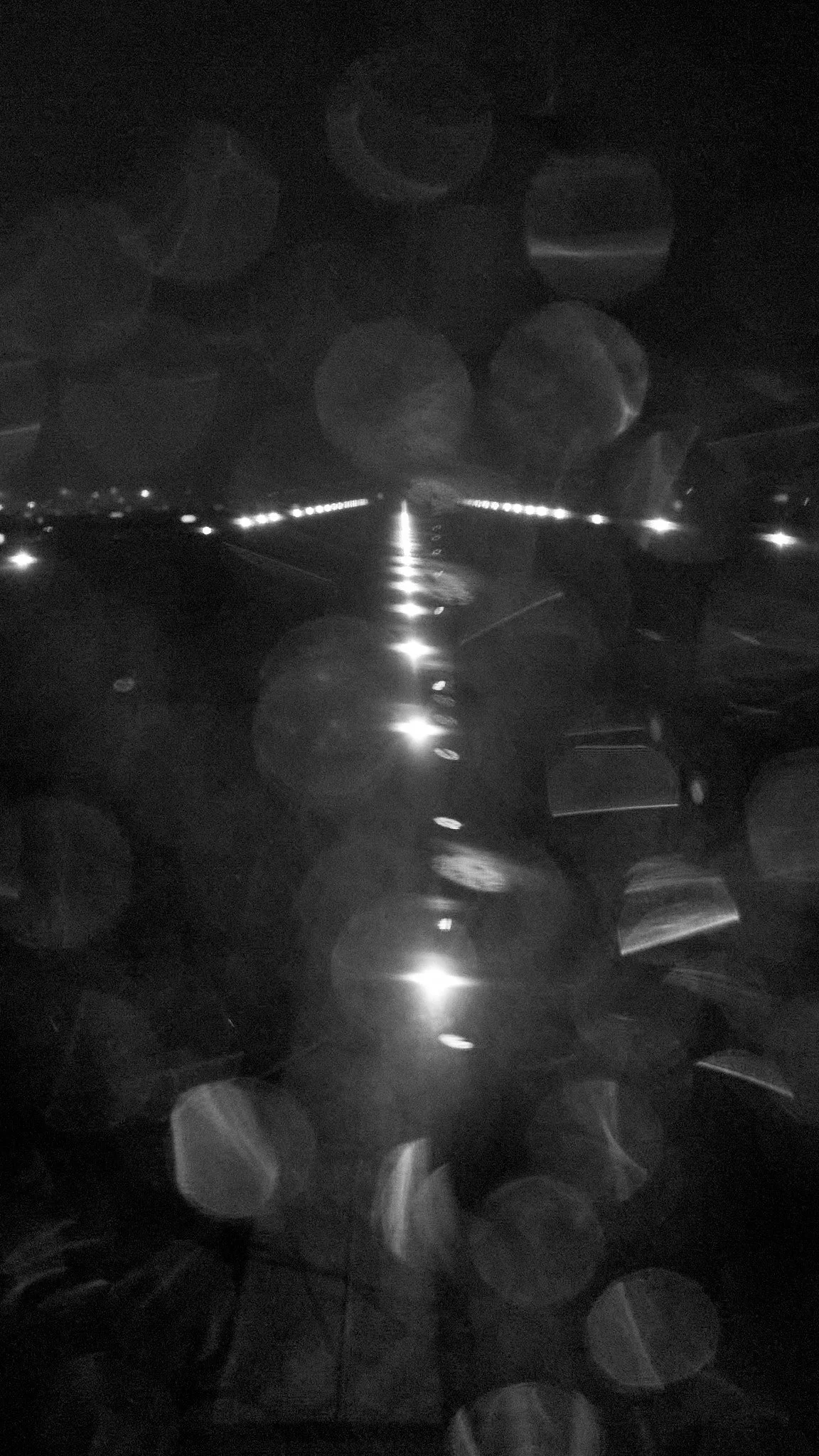 Nighttime view of a road with bright streetlights and reflective glare, seen through a rain-covered window with raindrops