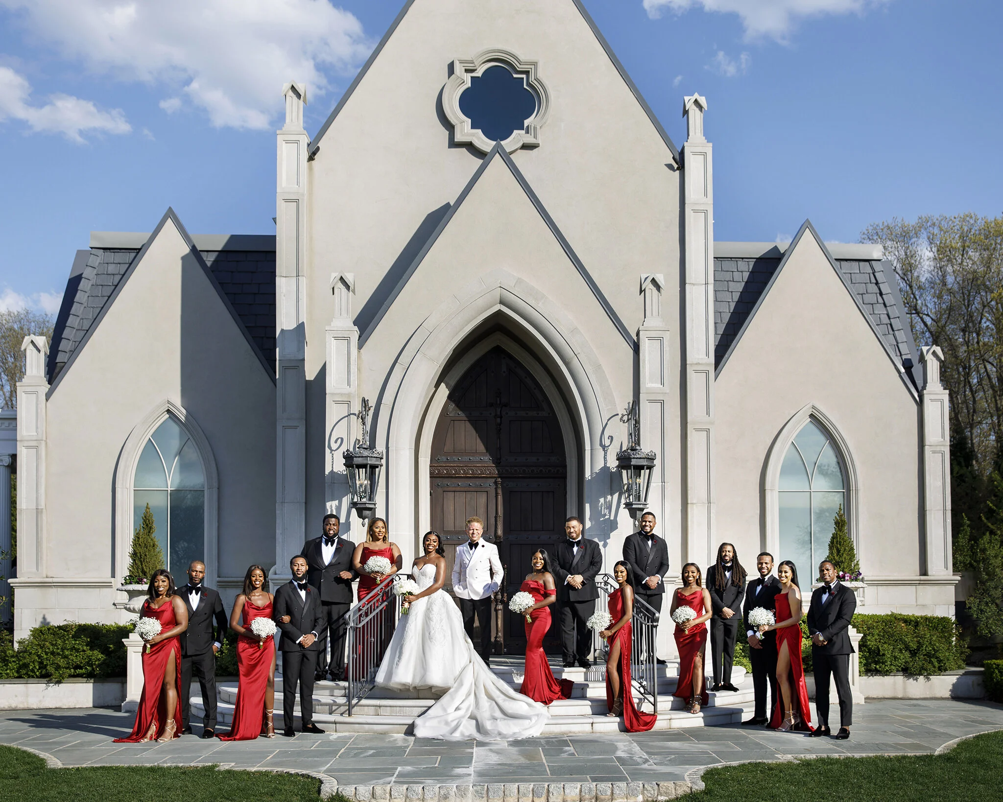 A wedding party standing on the front steps of a church. The bride in a white gown and the groom in a white tuxedo jacket are in the center. Bridesmaids in red dresses and groomsmen in black tuxedos are on either side. The church is a white stone bui