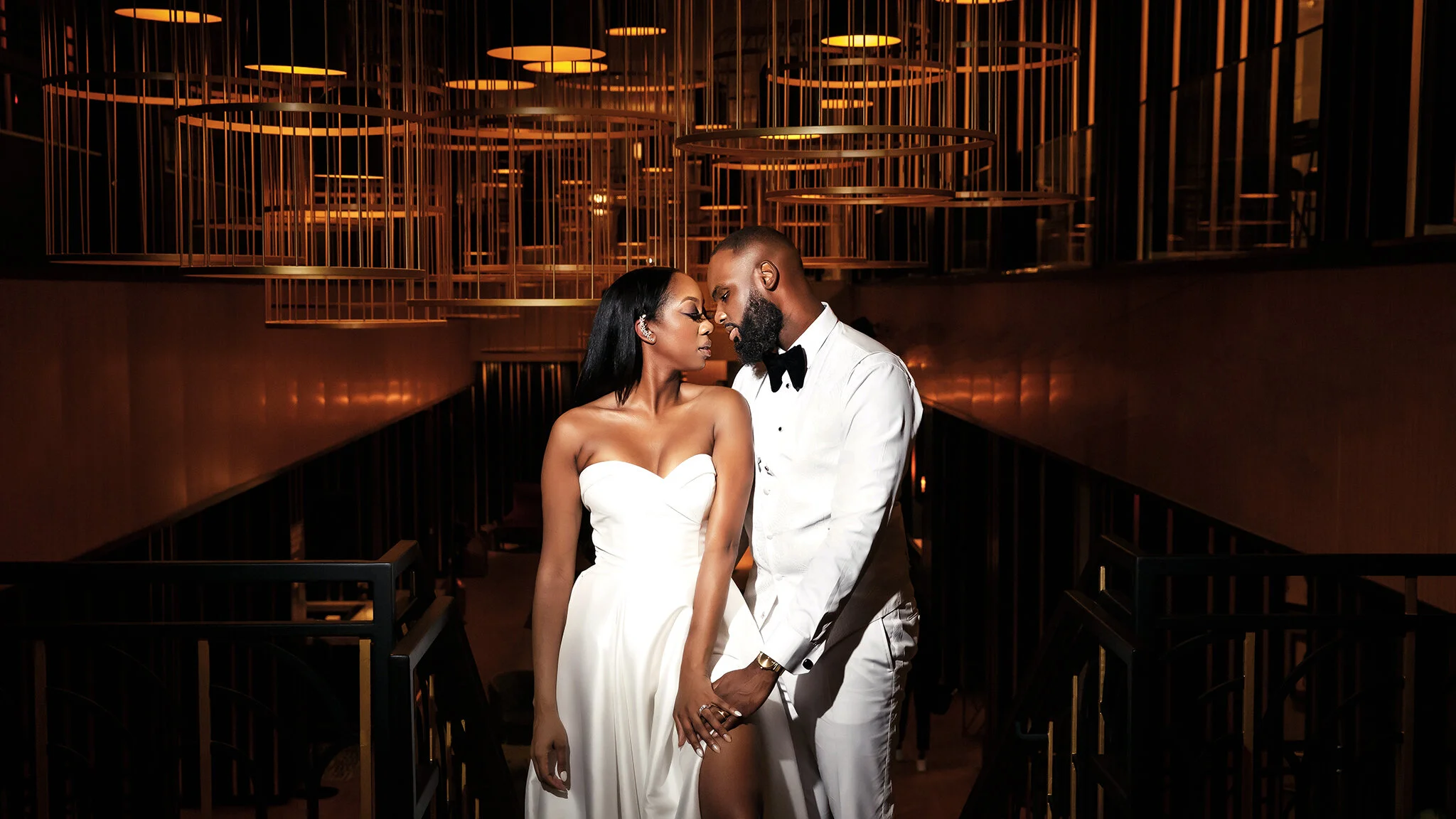 A couple in wedding attire in an elegant, dimly lit venue with modern gold circular chandeliers overhead. The woman is in a strapless white gown and the man is in a white tuxedo with a black bow tie, standing close and touching hands.